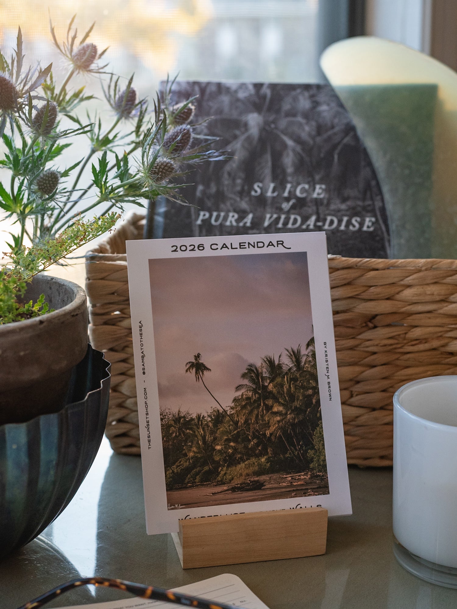 A 2026 Costa Rica calendar featuring a tropical beach scene with palm trees, displayed on a wooden stand on a tabletop. The calendar is styled in a home setting with a wicker basket, a potted plant, a candle, and a book in the background.