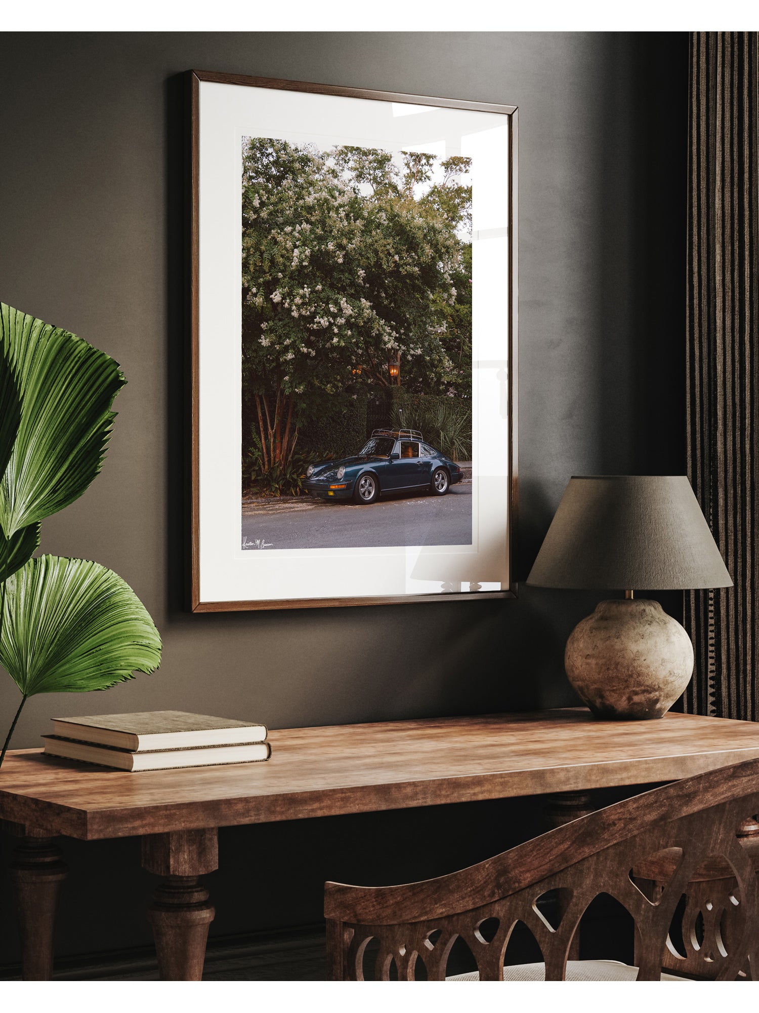 A framed art print featuring a vintage Porsche 911, displayed on a dark gray wall above a wooden console table with a lamp and books. Tucked beneath the cascading white blooms of a towering crepe myrtle, this vintage Porsche 911 awaits its next adventure. So what are you waiting for? Hop on in and channel that carefree feeling of adventure and speed with this air cooled vintage beauty, "911 Meeting". Vintage Porsche 911 photo print by Kristen M. Brown of Samba to the Sea for The Sunset Shop.