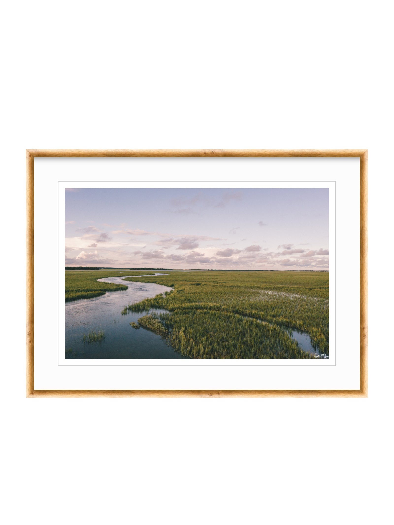 A framed landscape photograph of a winding river through green marshlands under a cloudy sky, presented in a light wood frame with a white mat on a white background. A scenic landscape photograph of a winding river flowing through a vast green marsh under a pale blue and pink sky. "Creek Serenade" photo print of golden hour over the a marsh wandering creek during high tide in Charleston, SC. Photo by Kristen M. Brown of Samba to the Sea for The Sunset Shop.