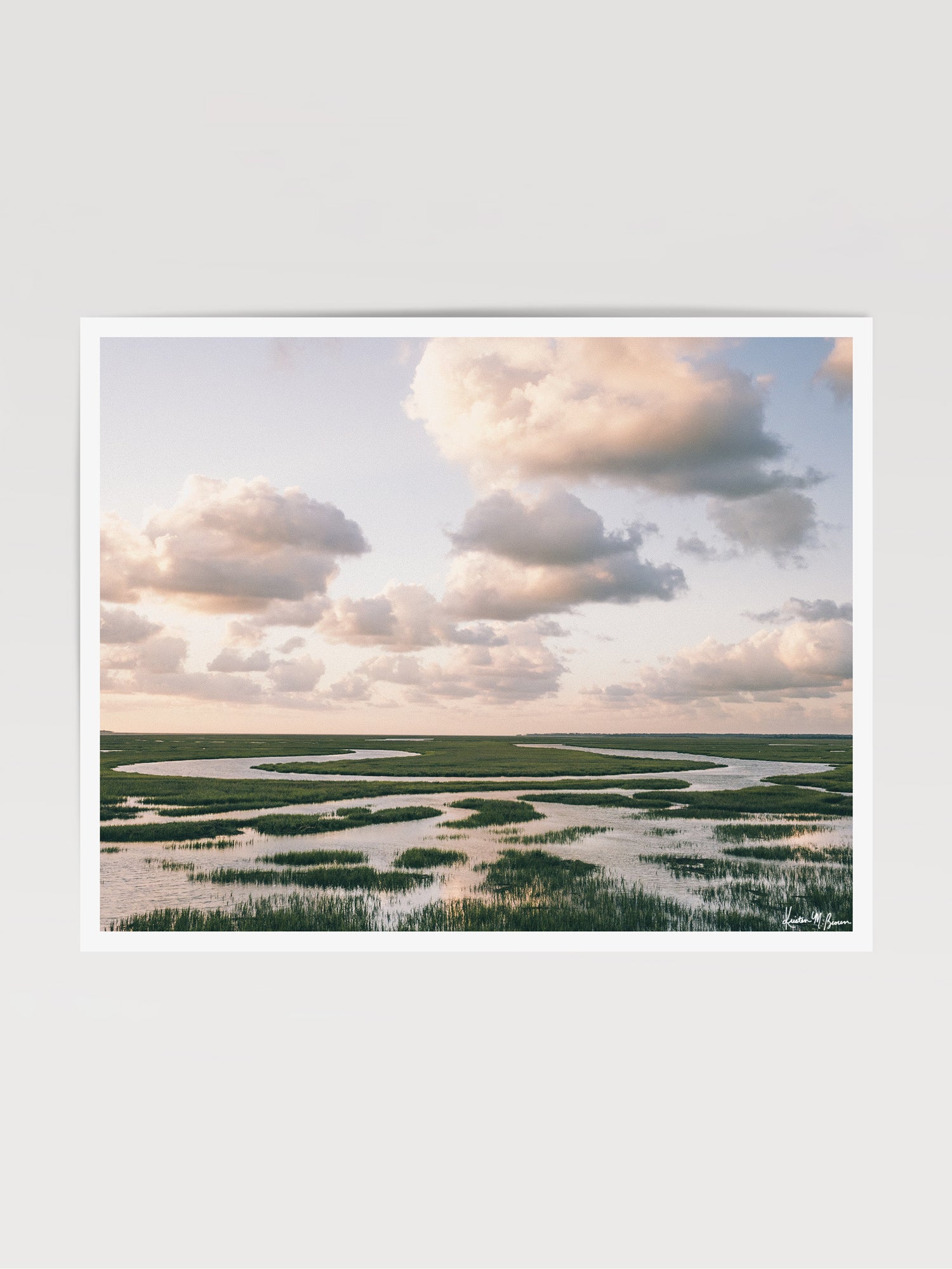 Landscape photograph of marsh with winding river and tall green marsh grass under a pastel sunrise sky filled with soft clouds. The Lowcountry breathes in endless curves of marsh grass and winding tidal creeks, where the horizon stretches as far as the eye can see. Across the vast coastal wetlands between Charleston and Savannah, you can follow the serpentining waterways as they carve through lush grasses. "Marsh Meander" photo print by Kristen M. Brown of Samba to the Sea for The Sunset Shop.