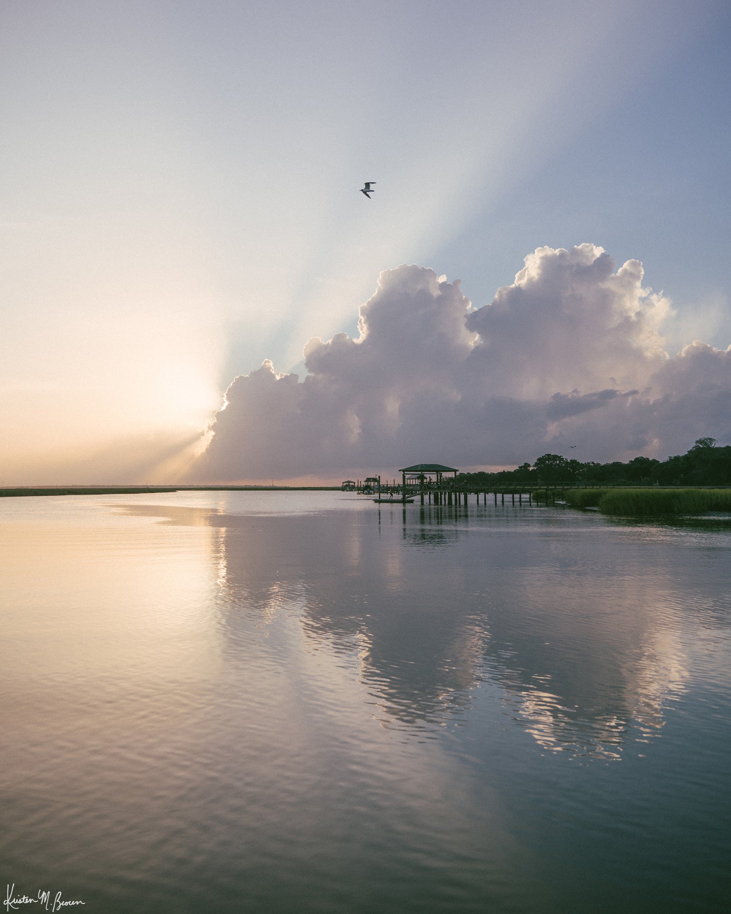 A scenic landscape photograph of a marsh during sunrise with sun rays breaking through clouds, a small bird in the sky, and wooden docks extending into the water. The scene is reflected clearly in the still water, with green marsh grass visible along the right shoreline. “Morning Glory” fine art photography print photographed by Kristen M. Brown, Samba to the Sea.