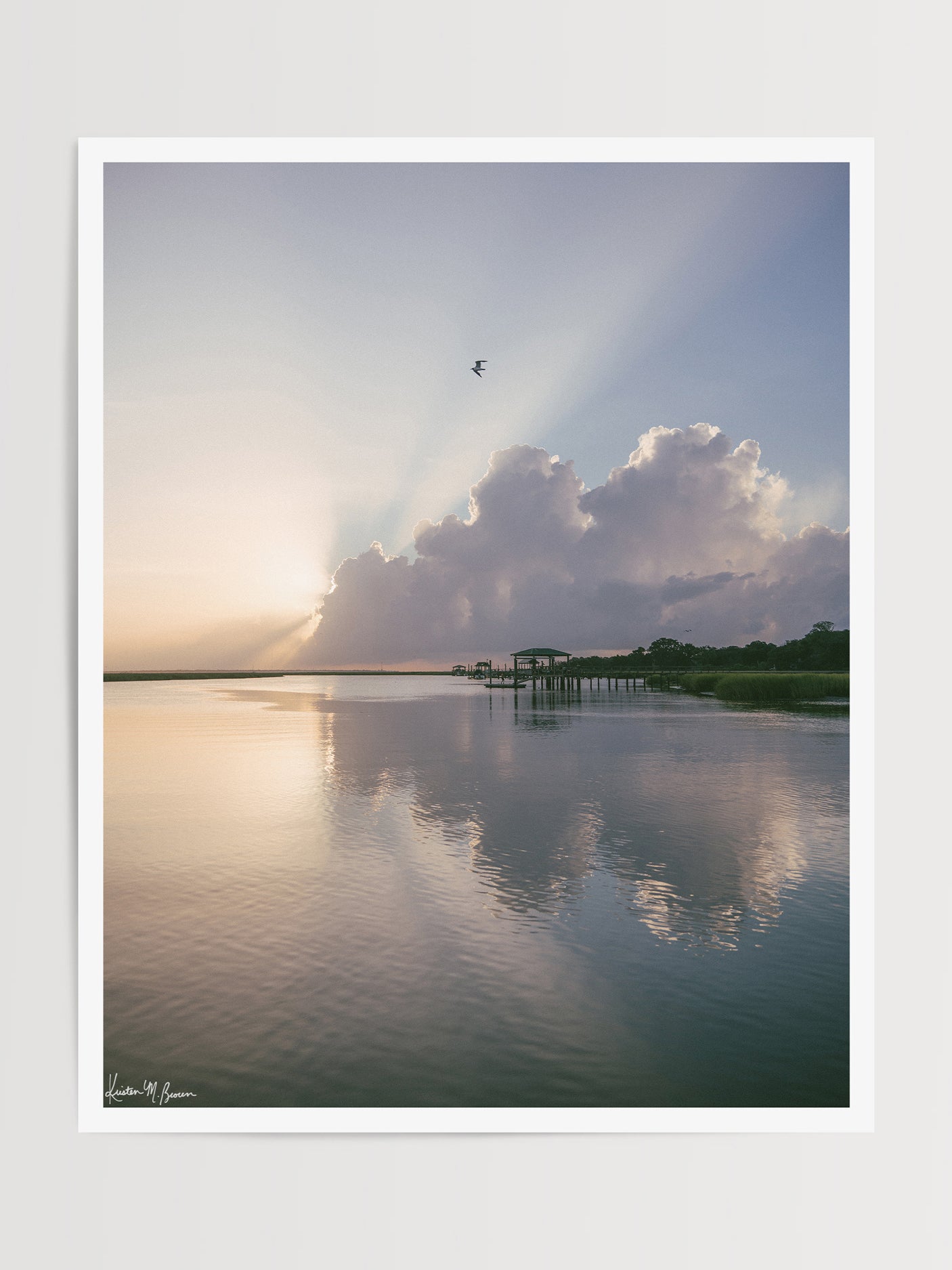 A scenic landscape photograph of a marsh during sunrise with sun rays breaking through clouds, a small bird in the sky, and wooden docks extending into the water. The scene is reflected clearly in the still water, with green marsh grass visible along the right shoreline. “Morning Glory” fine art photography print photographed by Kristen M. Brown, Samba to the Sea.