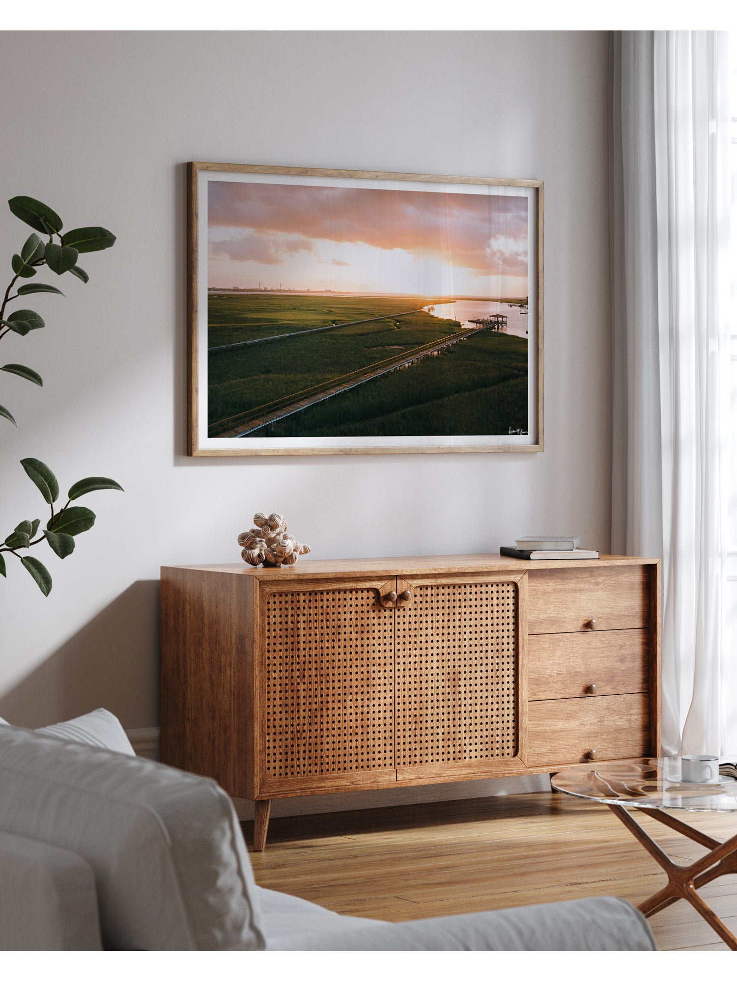 Aerial photograph of pastel pink sunrise over Charleston harbor and marsh with a boardwalk path, waterway, and view of downtown Charleston in the background. "Morning Harbor View" fine art photography print photographed by Kristen M. Brown, Samba to the Sea. Frame print displayed on a light gray wall above a wooden credenza in a modern living room setting.