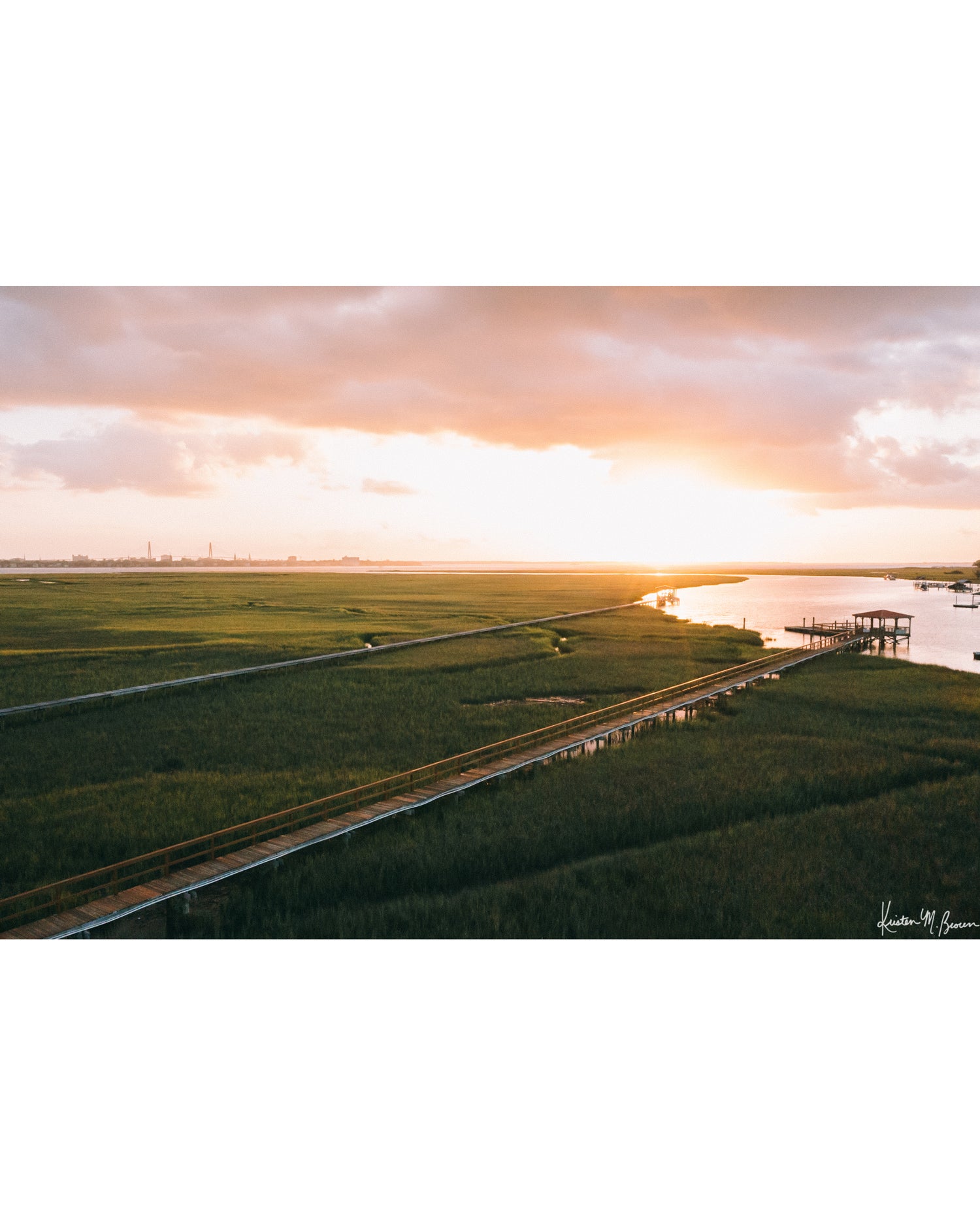 Aerial photograph of pastel pink sunrise over Charleston harbor and marsh with a boardwalk path, waterway, and view of downtown Charleston in the background. "Morning Harbor View" fine art photography print photographed by Kristen M. Brown, Samba to the Sea.