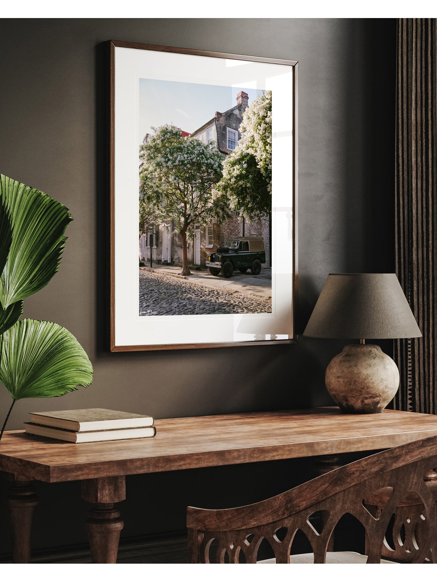 A framed vintage Land Rover photo print, displayed on a dark gray wall above a wooden console table with a lamp and books. A vintage green Land Rover Series truck British Army version with tan canvas top, parked on a cobblestone street in historic Charleston with blooming white Crepe Myrtle flowers. “Rover Blooms” is a journey into the charming streets of historic Charleston, where beauty and nostalgia meet. Vintage Land Rover photo print by Kristen M. Brown of Samba to the Sea for The Sunset Shop.