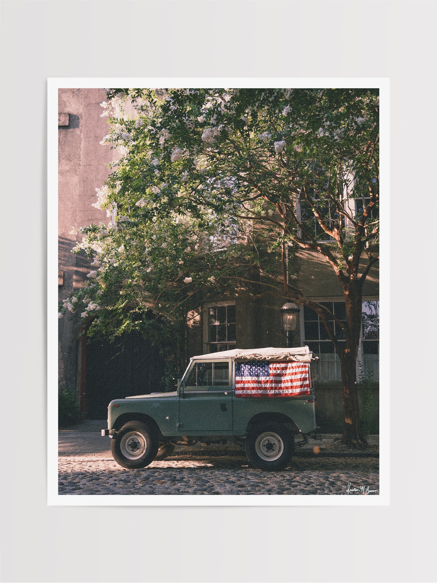 A vintage green Land Rover Series with an American flag draped over the rear window, parked on a cobblestone street in historic Charleston with blooming white Crepe Myrtle flowers. "Stars & Stripes & Series" is a journey into the charming streets of historic Charleston, where beauty and nostalgia meet. Vintage Land Rover photo print by Kristen M. Brown of Samba to the Sea for The Sunset Shop.