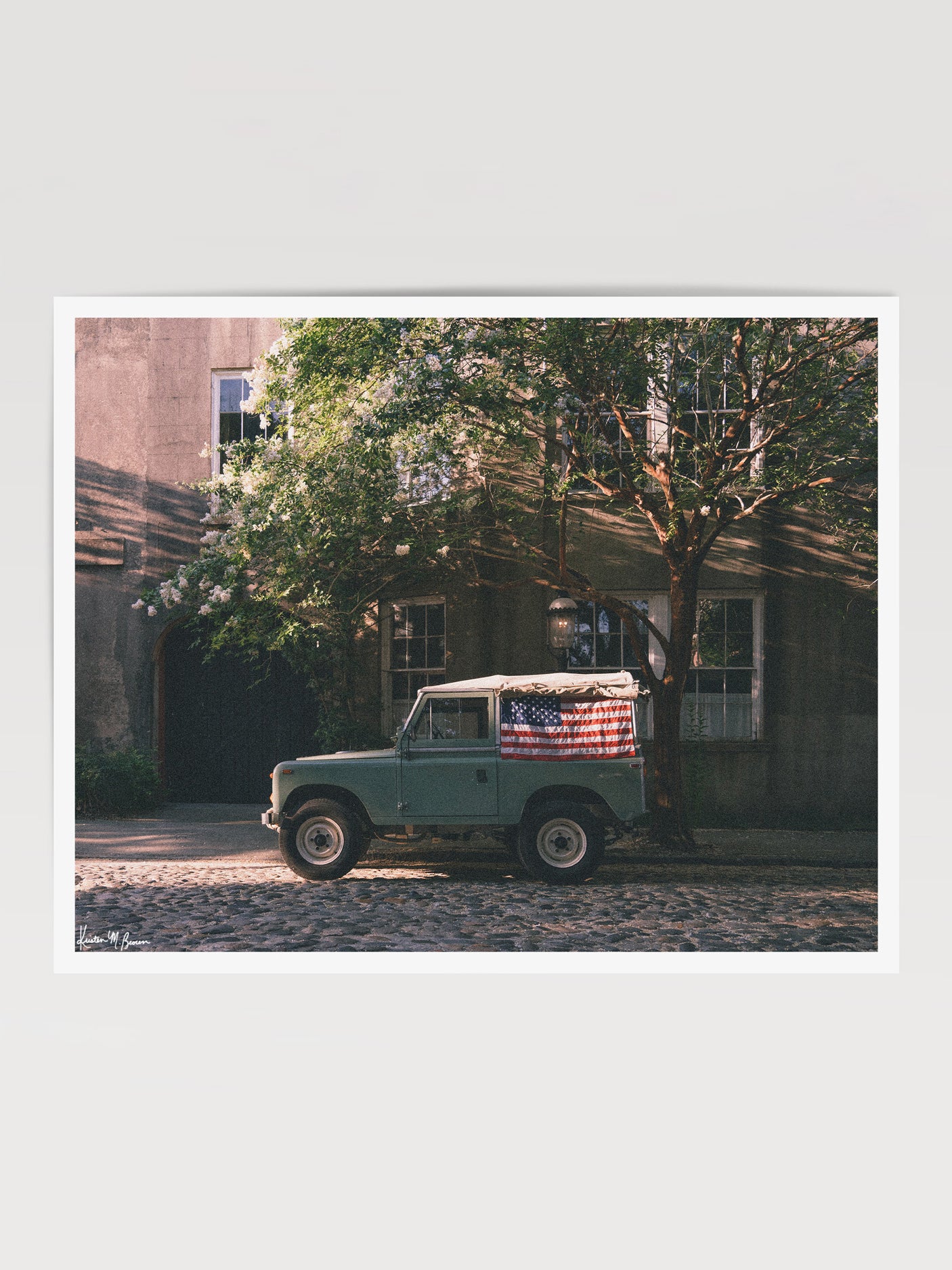 A vintage green Land Rover Series with an American flag draped over the rear window, parked on a cobblestone street in historic Charleston with blooming white Crepe Myrtle flowers. "Stars & Stripes & Series" is a journey into the charming streets of historic Charleston, where beauty and nostalgia meet. Vintage Land Rover photo print by Kristen M. Brown of Samba to the Sea for The Sunset Shop.