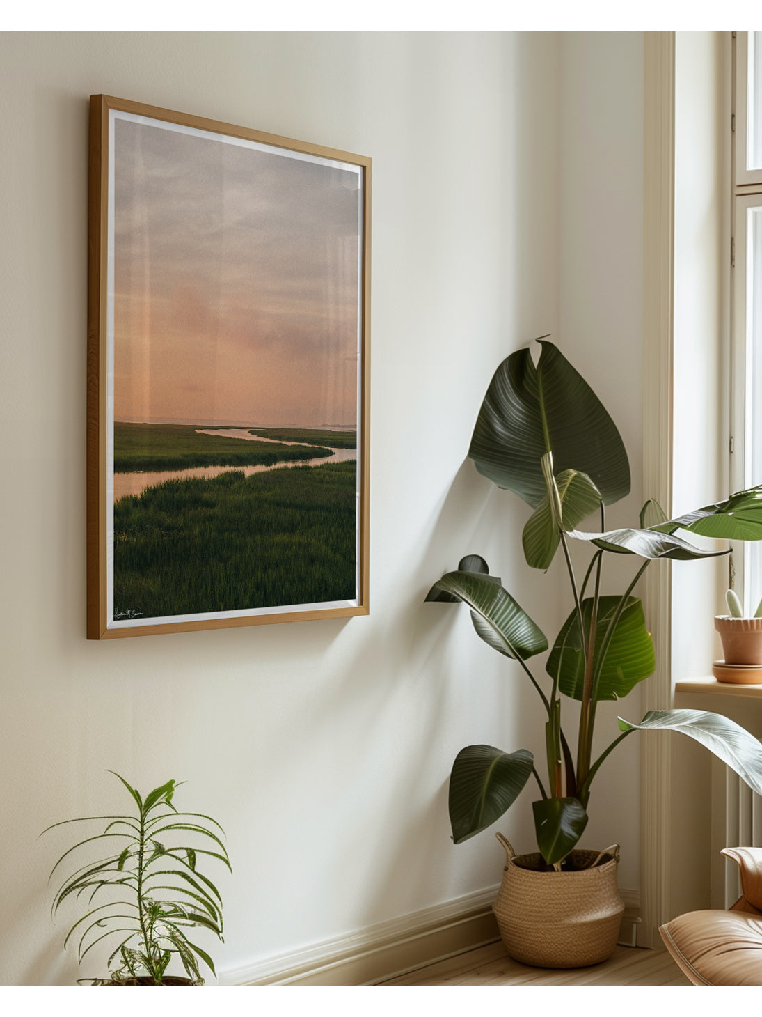 A scenic landscape photograph of a winding creek flowing through the marsh during sunrise, with the soft peach and pink clouds in the sky above and reflecting in the waters. "Tidal Glow" fine art photography print photographed by Kristen M. Brown, Samba to the Sea. Framed landscape art print styled on a cream wall in a bright room next to potted green plants.