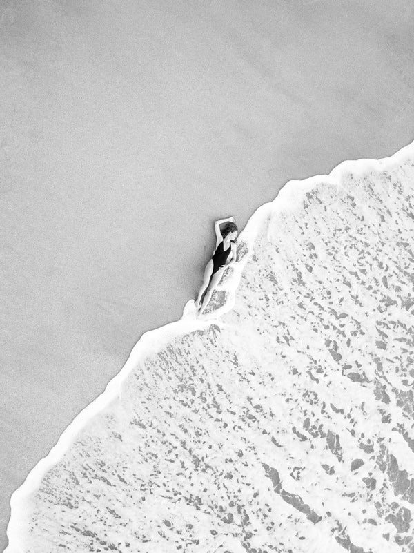Black and white image of woman laying on the beach as the waves reach her feet in Costa Rica. Photographed by Samba to the Sea for The Sunset Shop.
