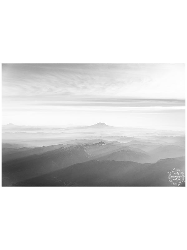 Black and white aerial image of late afternoon sun rays over Mt Rainier in Washington. What a glorious farewell the late afternoon sun gives the mountains. Magical late afternoon sun rays falling over Mt Rainier and the Cascade mountain range in Washington.