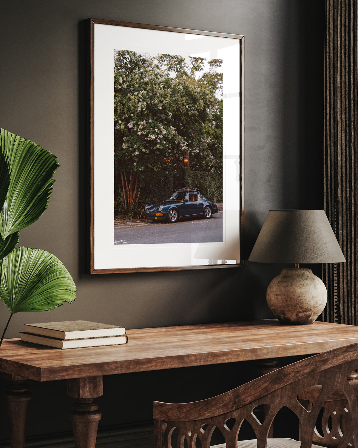 A framed art print featuring a vintage Porsche 911, displayed on a dark gray wall above a wooden console table with a lamp and books. Tucked beneath the cascading white blooms of a towering crepe myrtle, this vintage Porsche 911 awaits its next adventure. So what are you waiting for? Hop on in and channel that carefree feeling of adventure and speed with this air cooled vintage beauty, "911 Meeting".  Vintage Porsche 911 photo print by Kristen M. Brown of Samba to the Sea for The Sunset Shop.