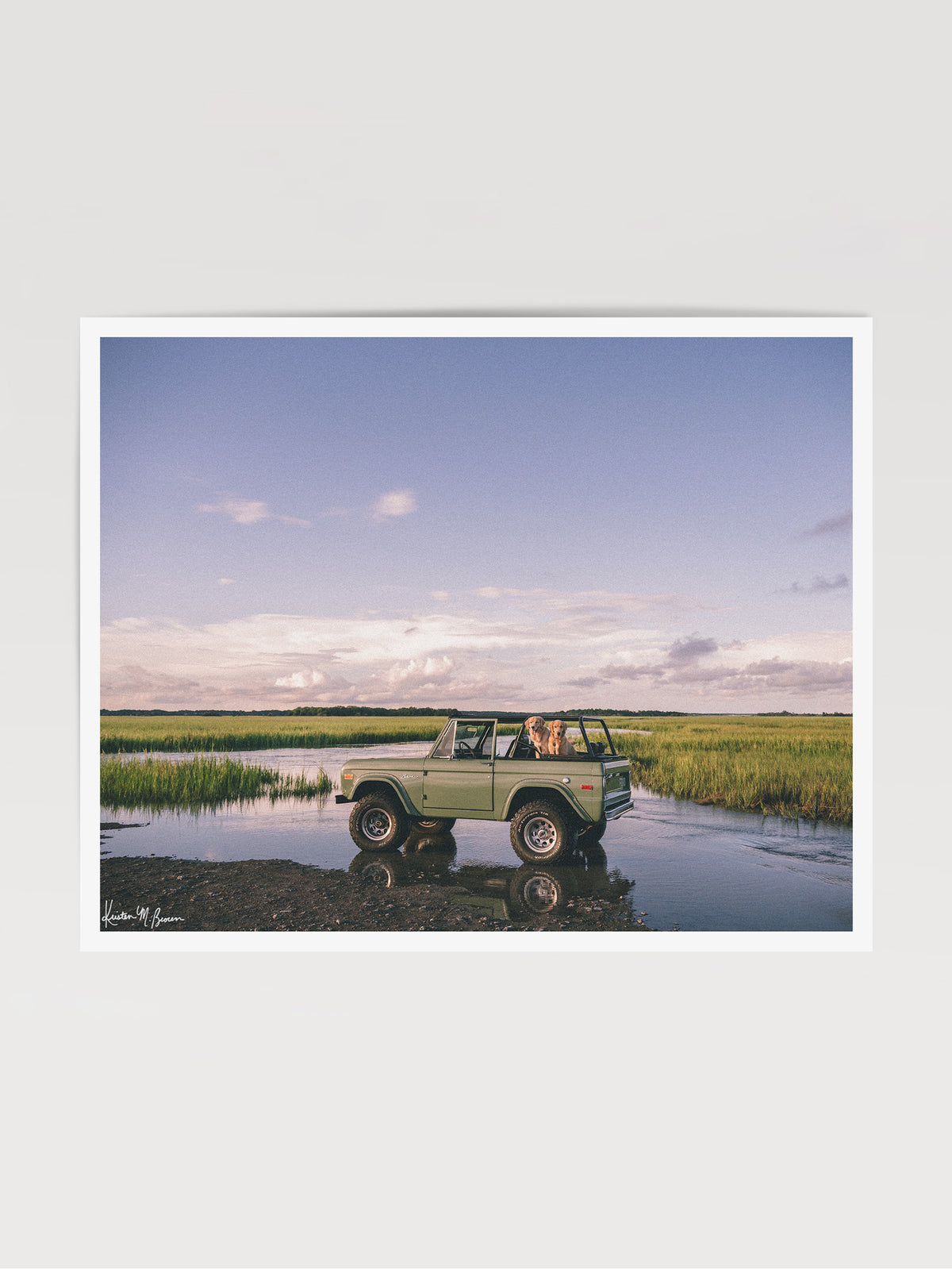 Photographic art print featuring a vintage green Ford Bronco along the marsh landscape under a pale blue sky with golden retrievers dogs waiting for an adventure. Lowcountry golden hours hit different when you’ve got four-wheel drive, a tailgate full of tail wags, and nowhere you’d rather be. "Best Friends and Bronco Rides" photo print of golden retrievers waiting for you to hop in a vintage Bronco to start the afternoon adventure along the marsh in Charleston, SC. Photograph by Kristen M. Brown.