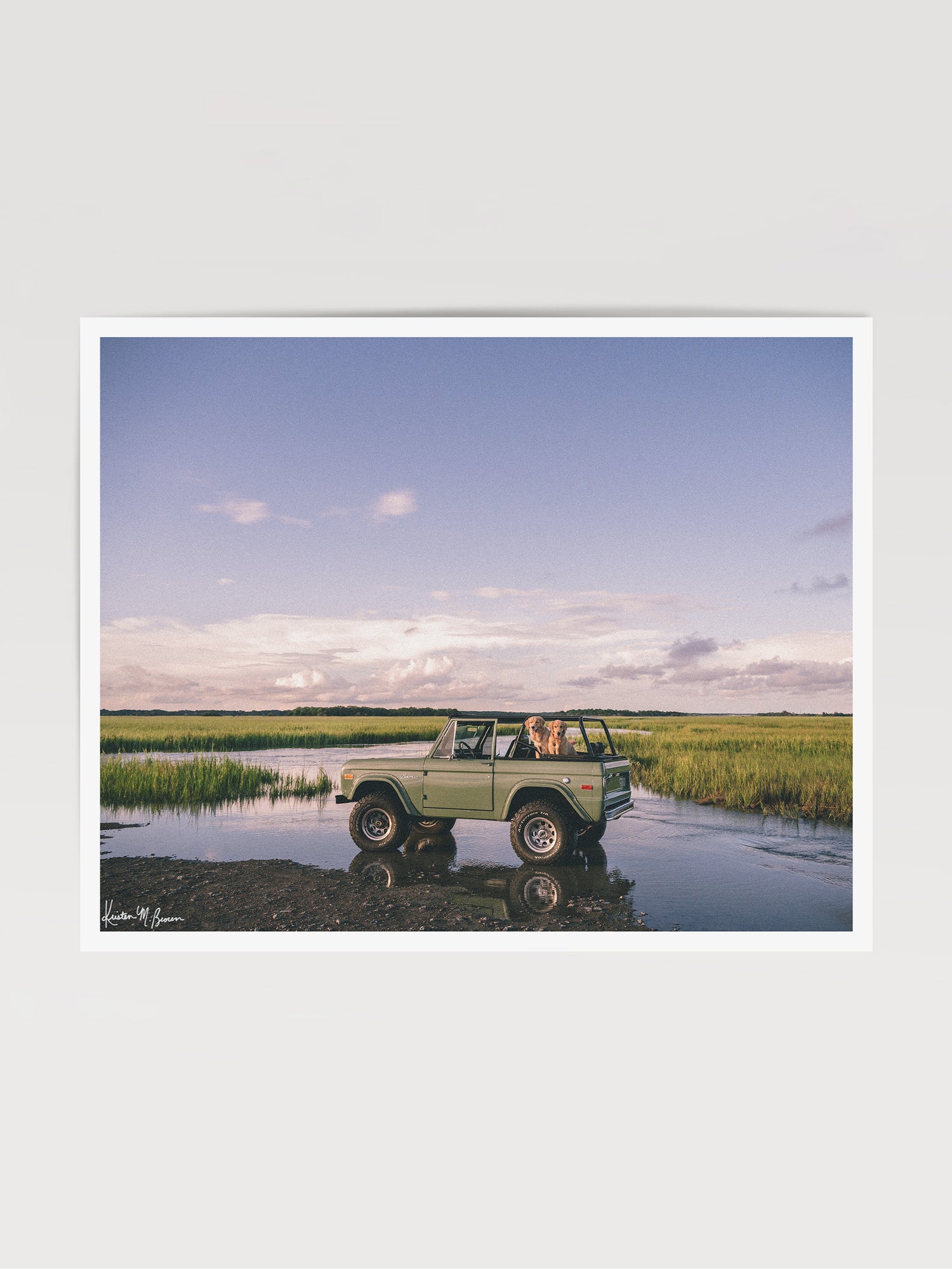 Photographic art print featuring a vintage green Ford Bronco along the marsh landscape under a pale blue sky with golden retrievers dogs waiting for an adventure. Lowcountry golden hours hit different when you’ve got four-wheel drive, a tailgate full of tail wags, and nowhere you’d rather be. "Best Friends and Bronco Rides" photo print of golden retrievers waiting for you to hop in a vintage Bronco to start the afternoon adventure along the marsh in Charleston, SC. Photograph by Kristen M. Brown.