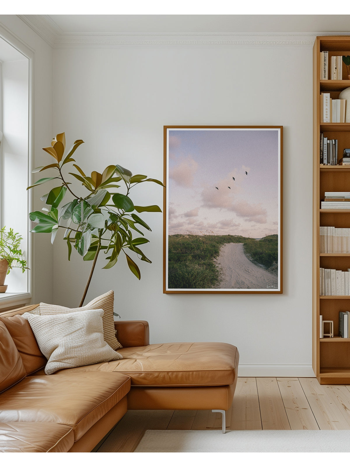 A framed beach path sunset art print with a tan leather sectional sofa, a tall potted plant, and a wooden bookshelf. Bring home that feeling of the warm ocean breeze flowing through your hair, the soft pastel evening light, and the gentle, timeless magic of the shoreline at day's end with "Cloud Patrol". Sunset beach photo print with soaring pelicans in Charleston, SC by Kristen M. Brown of Samba to the Sea for The Sunset Shop.