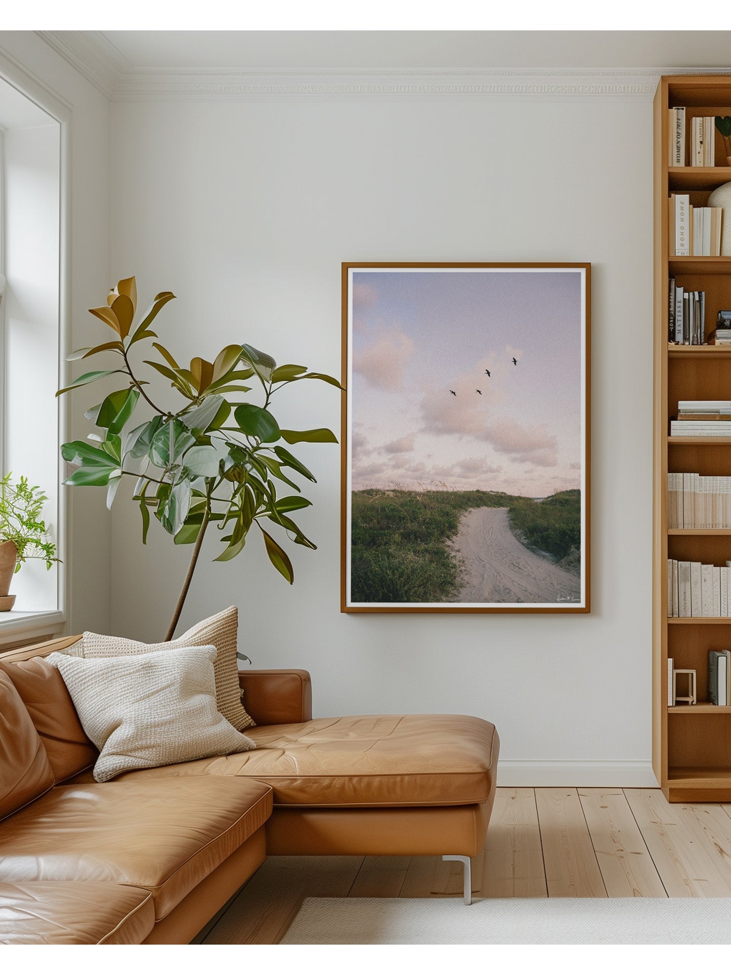 A framed beach path sunset art print with a tan leather sectional sofa, a tall potted plant, and a wooden bookshelf. Bring home that feeling of the warm ocean breeze flowing through your hair, the soft pastel evening light, and the gentle, timeless magic of the shoreline at day's end with "Cloud Patrol". Sunset beach photo print with soaring pelicans in Charleston, SC by Kristen M. Brown of Samba to the Sea for The Sunset Shop.