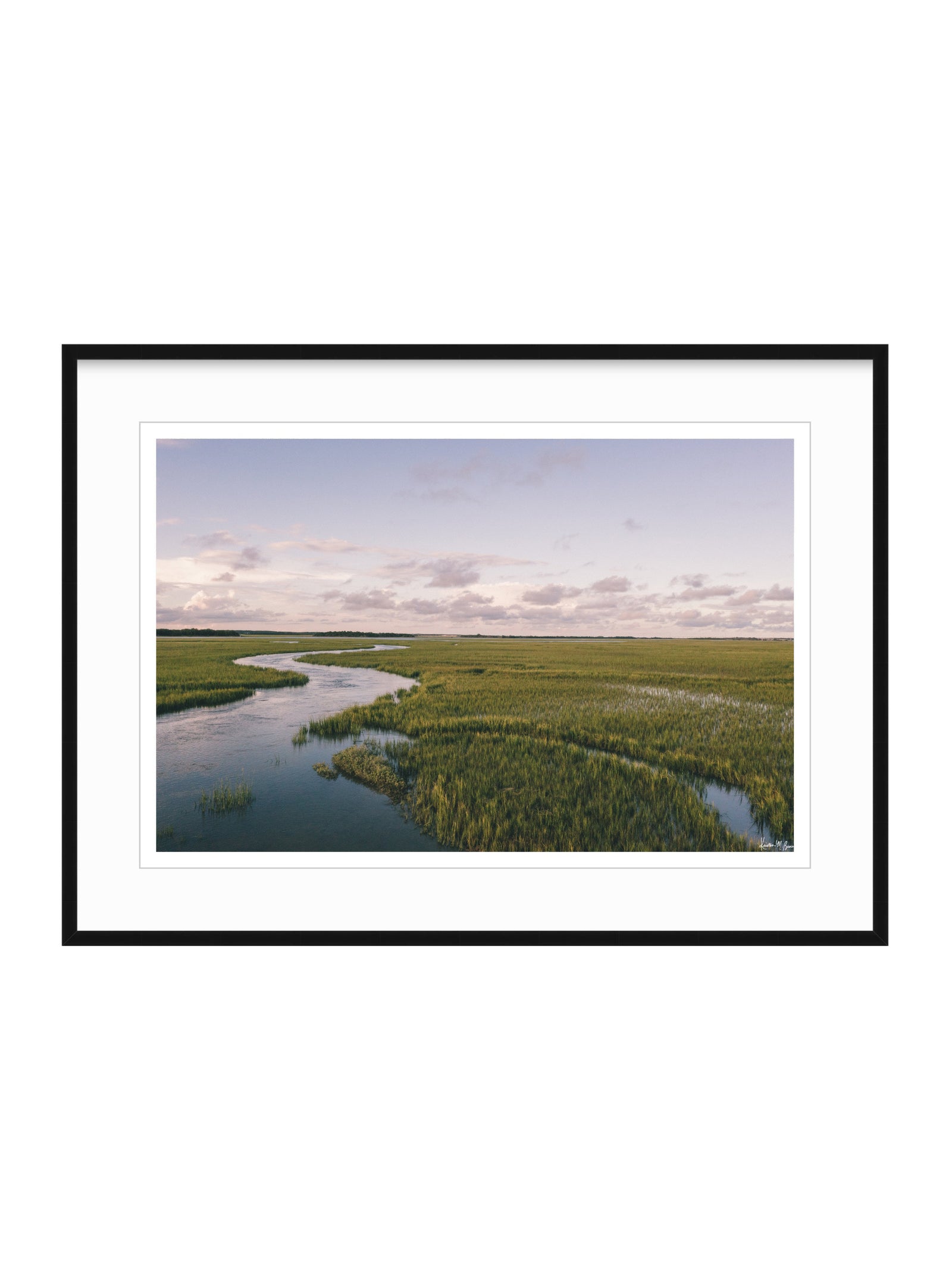 A framed landscape photograph of a winding river through green marshlands under a pale sky, presented with a white mat and black frame on a white background. A scenic landscape photograph of a winding river flowing through a vast green marsh under a pale blue and pink sky. "Creek Serenade" photo print of golden hour over the a marsh wandering creek during high tide in Charleston, SC. Photo by Kristen M. Brown of Samba to the Sea for The Sunset Shop.