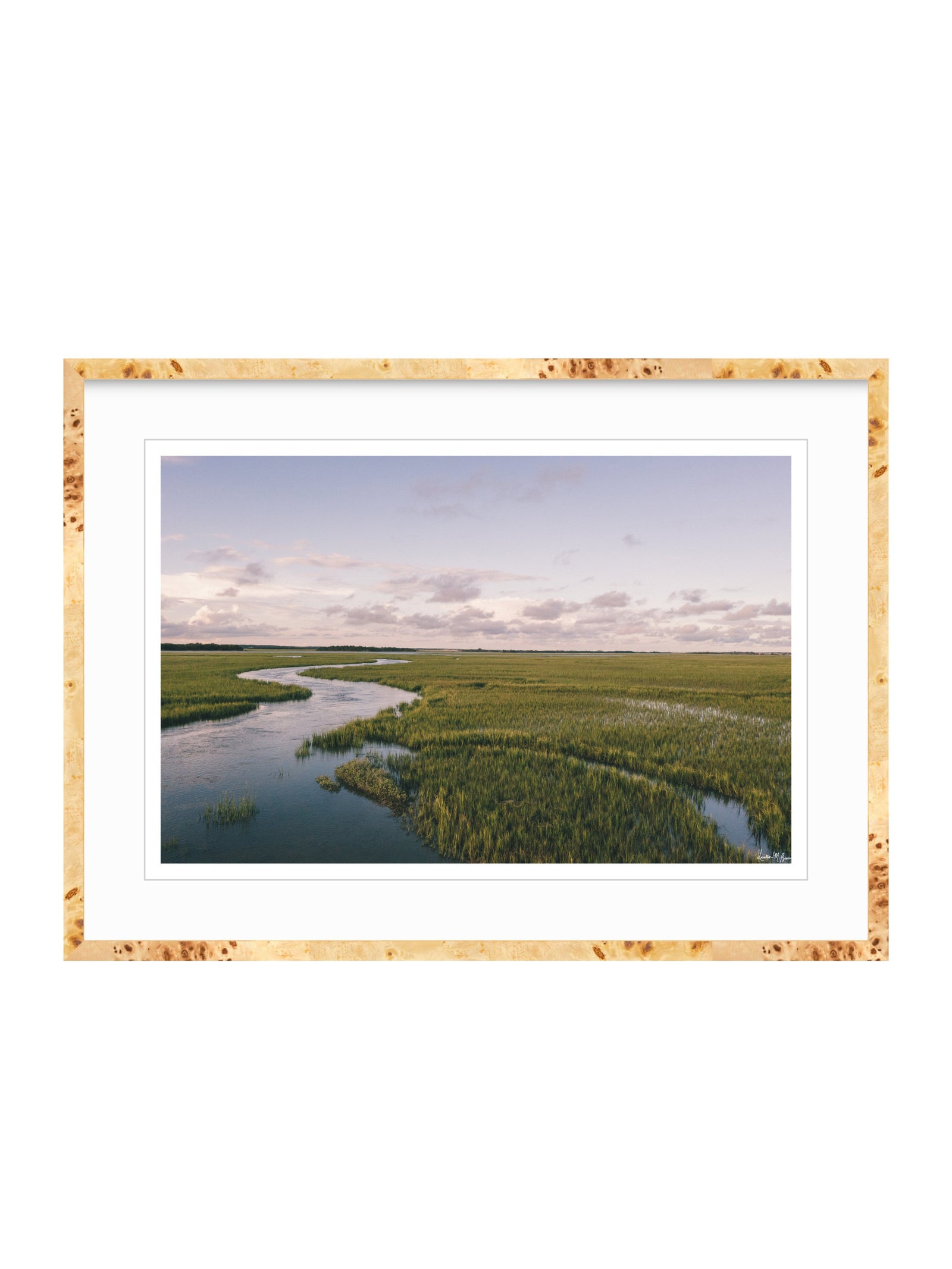 A framed landscape photograph of a winding river through green marshlands under a pale sky, presented in a burlwood frame with a white mat on a white background. A scenic landscape photograph of a winding river flowing through a vast green marsh under a pale blue and pink sky. "Creek Serenade" photo print of golden hour over the a marsh wandering creek during high tide in Charleston, SC. Photo by Kristen M. Brown of Samba to the Sea for The Sunset Shop.