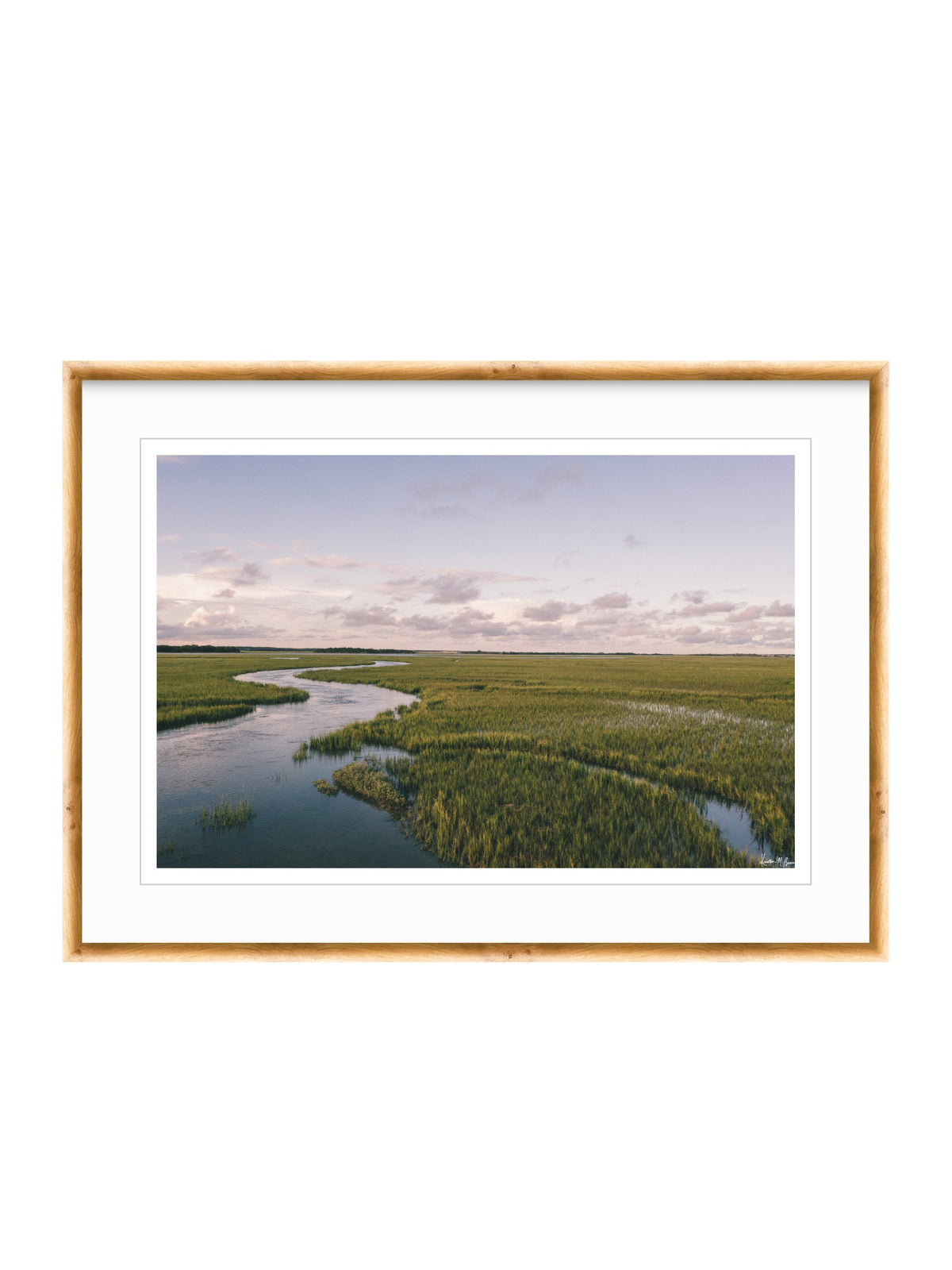 A framed landscape photograph of a winding river through green marshlands under a cloudy sky, presented in a light wood frame with a white mat on a white background. A scenic landscape photograph of a winding river flowing through a vast green marsh under a pale blue and pink sky. "Creek Serenade" photo print of golden hour over the a marsh wandering creek during high tide in Charleston, SC. Photo by Kristen M. Brown of Samba to the Sea for The Sunset Shop.