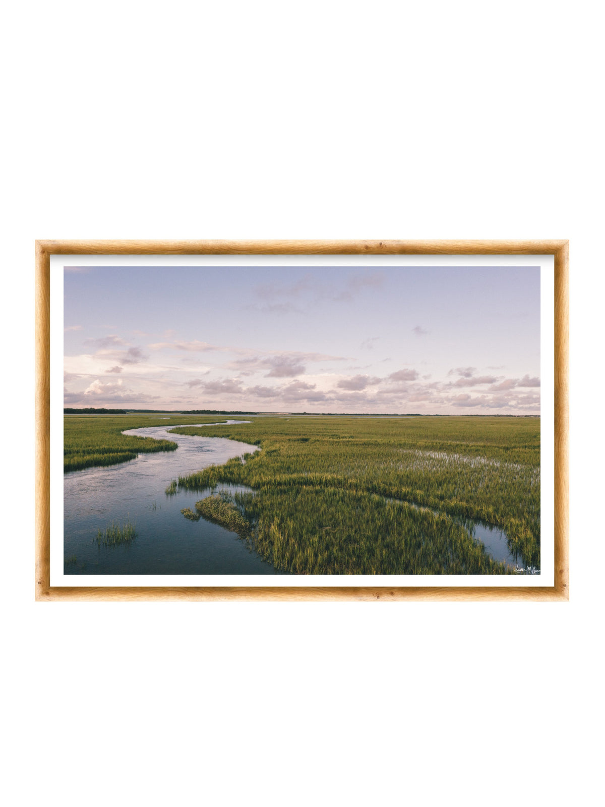 A framed landscape photograph of a winding river through green marshlands under a pale sky, presented in a light wood frame with a white mat on a white background. A scenic landscape photograph of a winding river flowing through a vast green marsh under a pale blue and pink sky. "Creek Serenade" photo print of golden hour over the a marsh wandering creek during high tide in Charleston, SC. Photo by Kristen M. Brown of Samba to the Sea for The Sunset Shop.