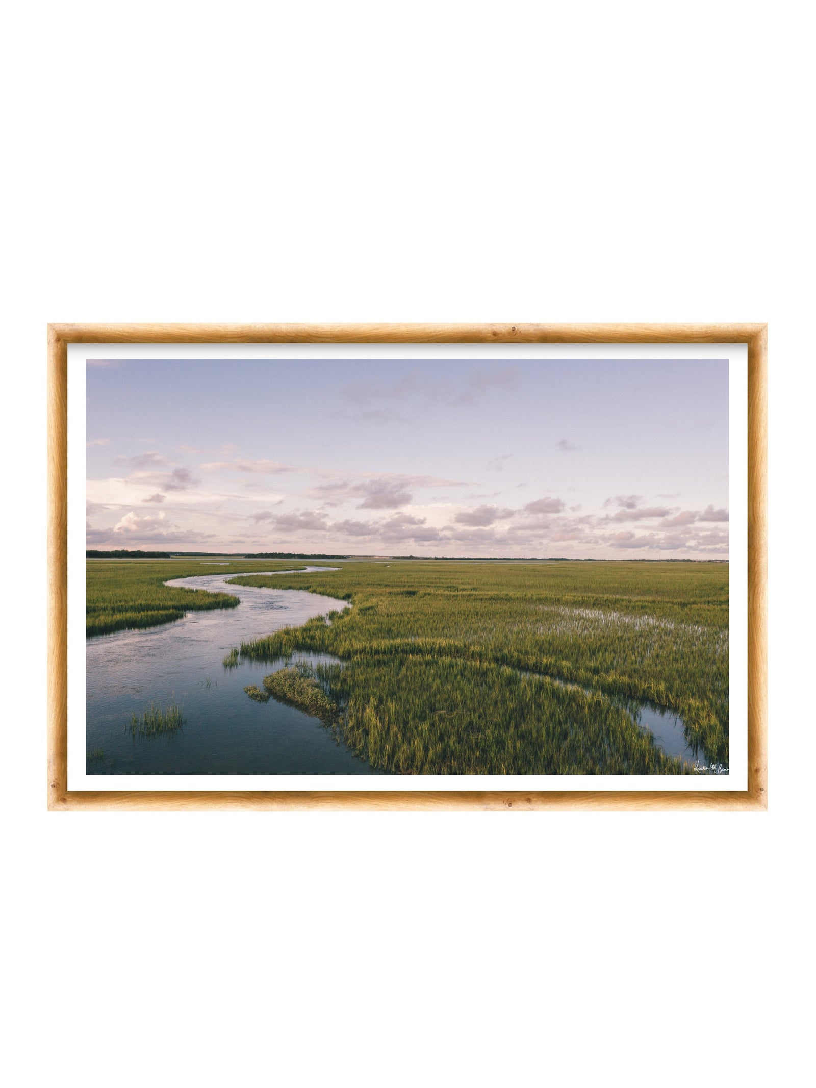 A framed landscape photograph of a winding river through green marshlands under a pale sky, presented in a light wood frame with a white mat on a white background. A scenic landscape photograph of a winding river flowing through a vast green marsh under a pale blue and pink sky. "Creek Serenade" photo print of golden hour over the a marsh wandering creek during high tide in Charleston, SC. Photo by Kristen M. Brown of Samba to the Sea for The Sunset Shop.