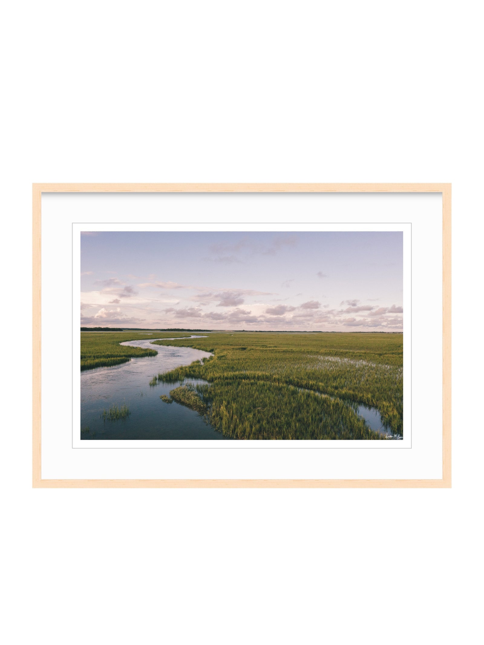 A framed landscape photograph of a winding river through green marshlands under a pale sky, presented in a light wood frame with a white mat on a white background. A scenic landscape photograph of a winding river flowing through a vast green marsh under a pale blue and pink sky. "Creek Serenade" photo print of golden hour over the a marsh wandering creek during high tide in Charleston, SC. Photo by Kristen M. Brown of Samba to the Sea for The Sunset Shop.