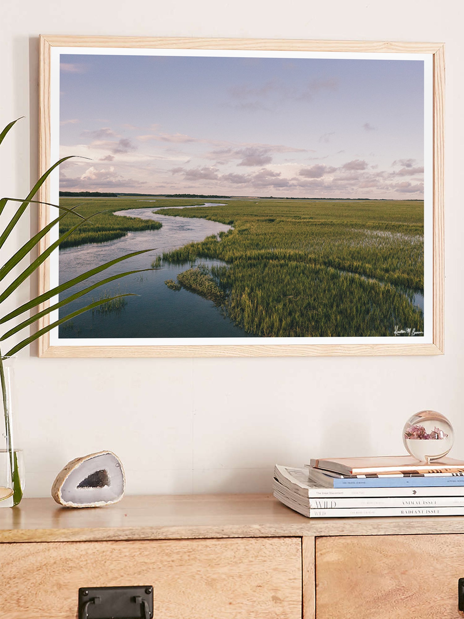 A framed landscape art print depicting a winding river through green marshlands under a cloudy sky, styled on a light wall above a wooden console table with books and a plant. "Creek Serenade" photo print of golden hour over the a marsh wandering creek during high tide in Charleston, SC. Photo by Kristen M. Brown of Samba to the Sea for The Sunset Shop.