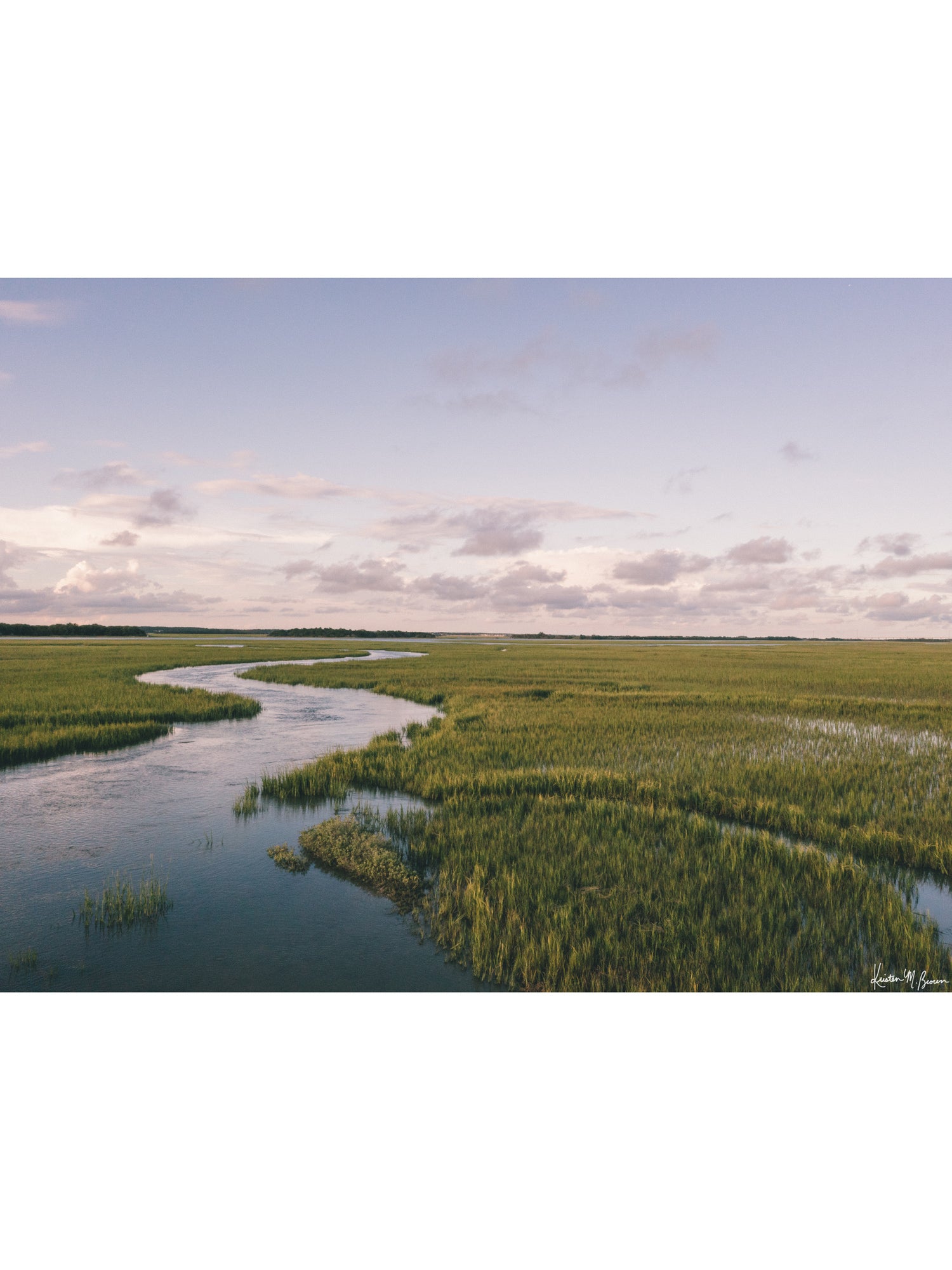A scenic landscape photograph of a winding river flowing through a vast green marsh under a pale blue and pink sky. "Creek Serenade" photo print of golden hour over the a marsh wandering creek during high tide in Charleston, SC. Photo by Kristen M. Brown of Samba to the Sea for The Sunset Shop.