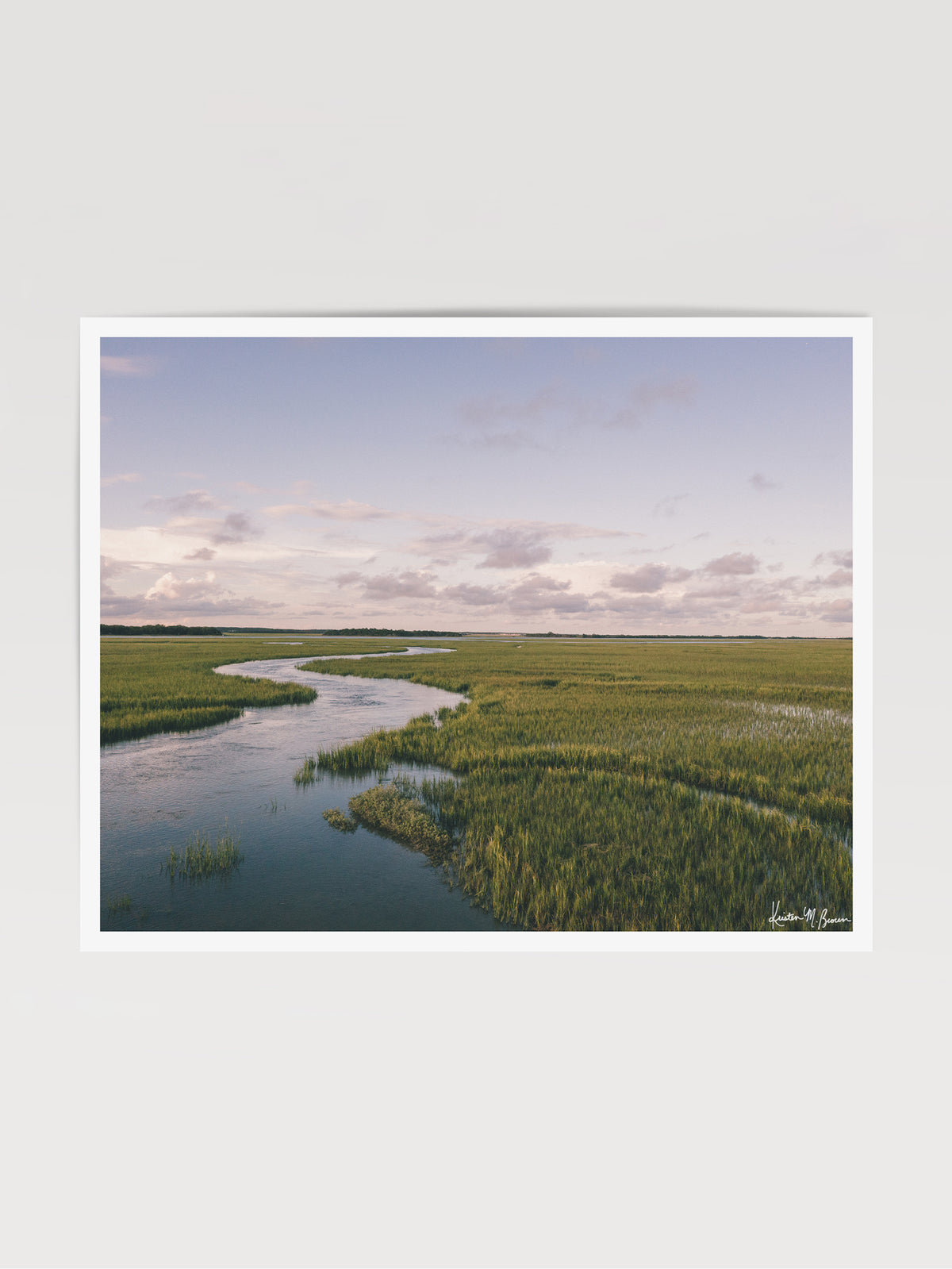 A scenic landscape photograph of a winding river flowing through a vast green marsh under a pale blue and pink sky. "Creek Serenade" photo print of golden hour over the a marsh wandering creek during high tide in Charleston, SC. Photo by Kristen M. Brown of Samba to the Sea for The Sunset Shop.