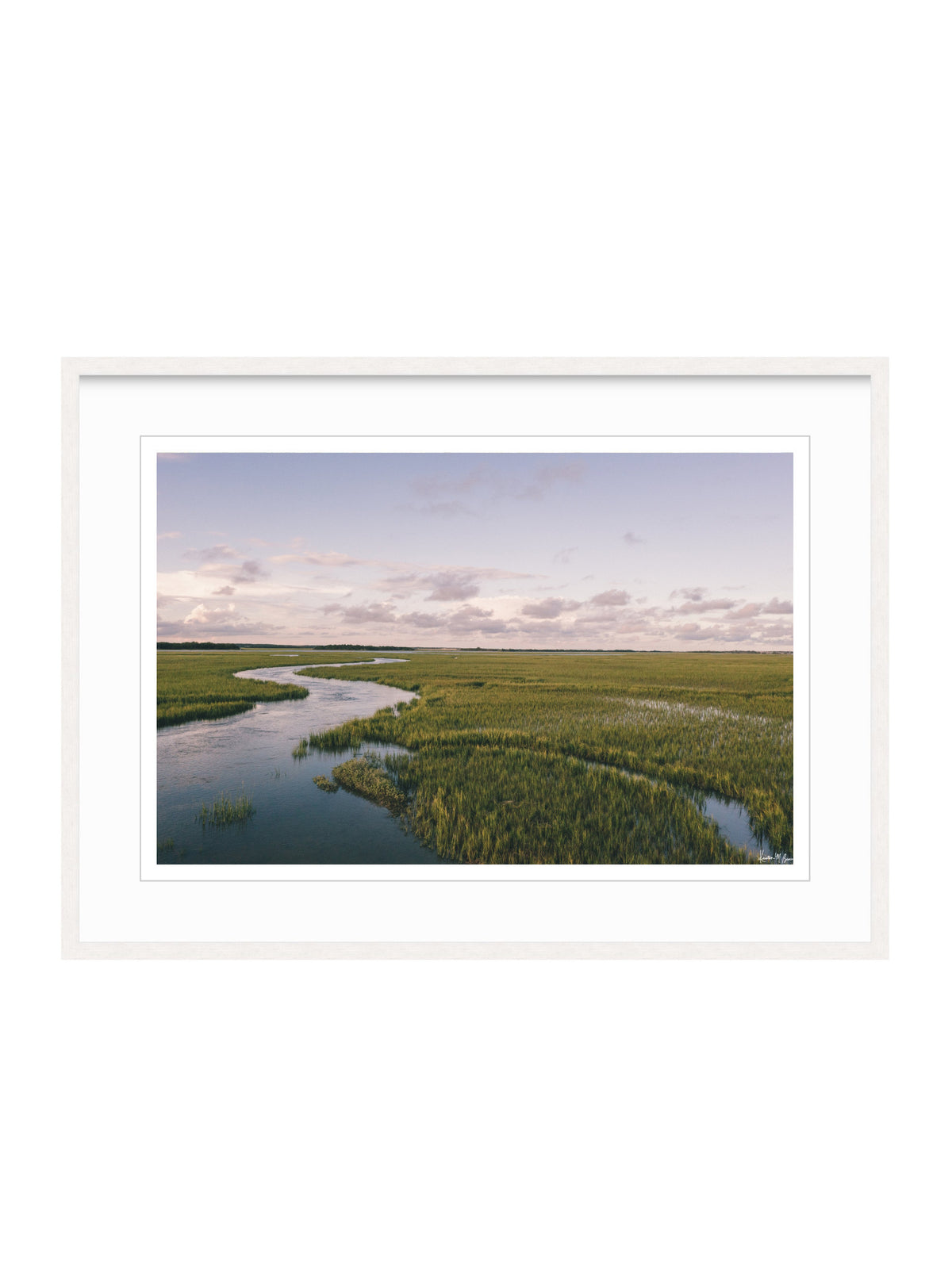 A framed landscape photograph of a winding river through green marshlands under a cloudy sky, presented in a white frame with a white mat on a white background. A scenic landscape photograph of a winding river flowing through a vast green marsh under a pale blue and pink sky. "Creek Serenade" photo print of golden hour over the a marsh wandering creek during high tide in Charleston, SC. Photo by Kristen M. Brown of Samba to the Sea for The Sunset Shop.