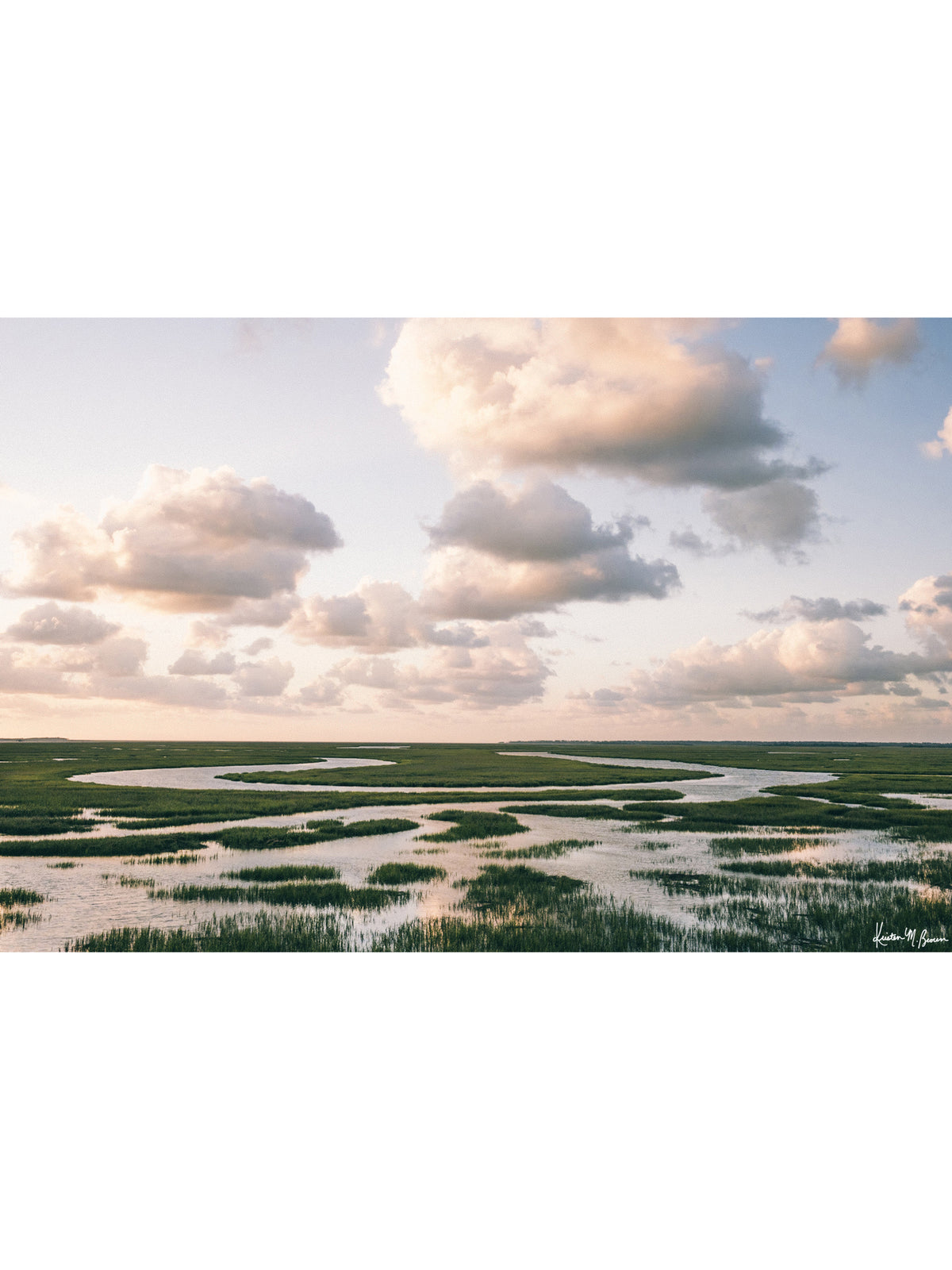 Landscape photograph of marsh with winding river and tall green marsh grass under a pastel sunrise sky filled with soft  clouds. The Lowcountry breathes in endless curves of marsh grass and winding tidal creeks, where the horizon stretches as far as the eye can see. Across the vast coastal wetlands between Charleston and Savannah, you can follow the serpentining waterways as they carve through lush grasses. "Marsh Meander" photo print by Kristen M. Brown of Samba to the Sea for The Sunset Shop.