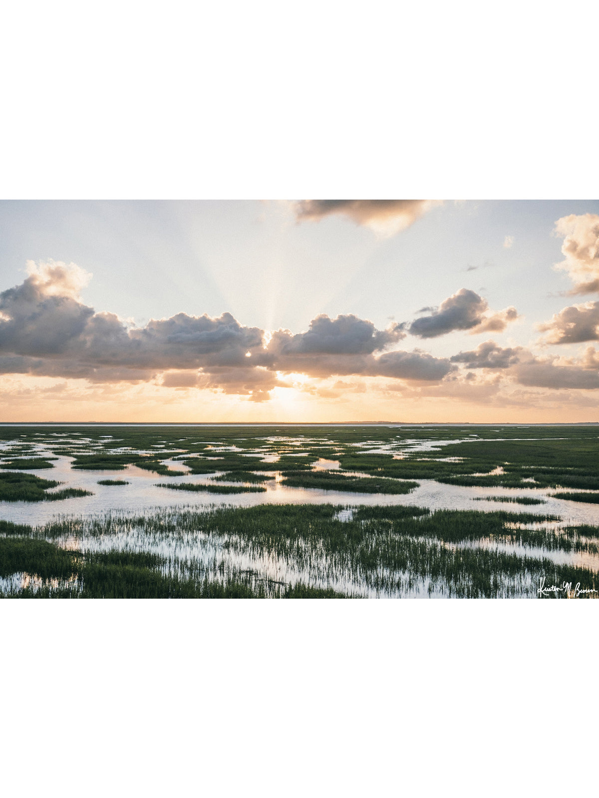 A scenic landscape photograph of a marsh with winding water channels and marsh grass under a dramatic sunrise sky with sun rays breaking through clouds. As the first rays of dawn beam through a crown of morning clouds, the Lowcountry marsh awakens in a breathtaking display of golden light and serene reflection. “Morning Beams” sunrise rays and reflections over the high tide marsh in the Lowcountry. Photo print by Kristen M. Brown of Samba to the Sea for The Sunset Shop.