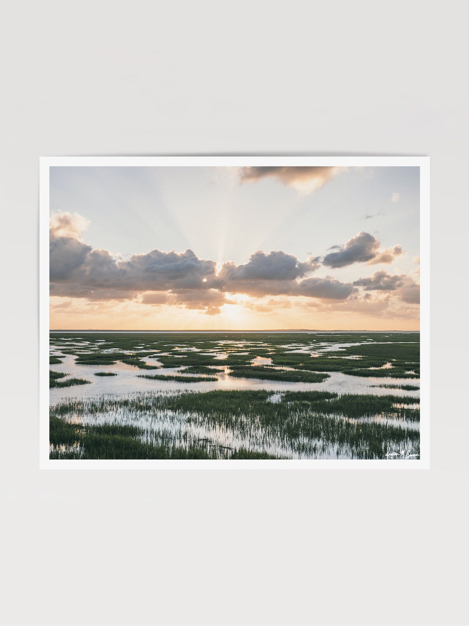 A scenic landscape photograph of a marsh with winding water channels and marsh grass under a dramatic sunrise sky with sun rays breaking through clouds. As the first rays of dawn beam through a crown of morning clouds, the Lowcountry marsh awakens in a breathtaking display of golden light and serene reflection. “Morning Beams” sunrise rays and reflections over the high tide marsh in the Lowcountry. Photo print by Kristen M. Brown of Samba to the Sea for The Sunset Shop.