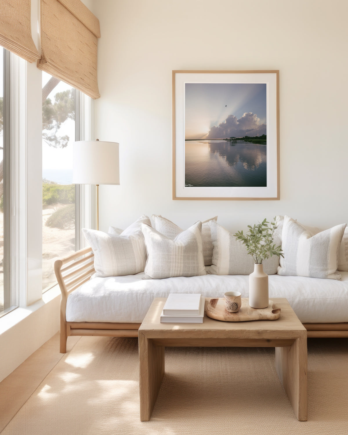 A framed scenic landscape photograph of a marsh during sunrise with sun rays breaking through clouds, a small bird in the sky, and wooden docks extending into the water. The scene is reflected clearly in the still water, with green marsh grass visible along the right shoreline. “Morning Glory” fine art photography print photographed by Kristen M. Brown, Samba to the Sea. Framed print hangs on a cream wall above a light wood sofa with white pillows in a bright, coastal living room.