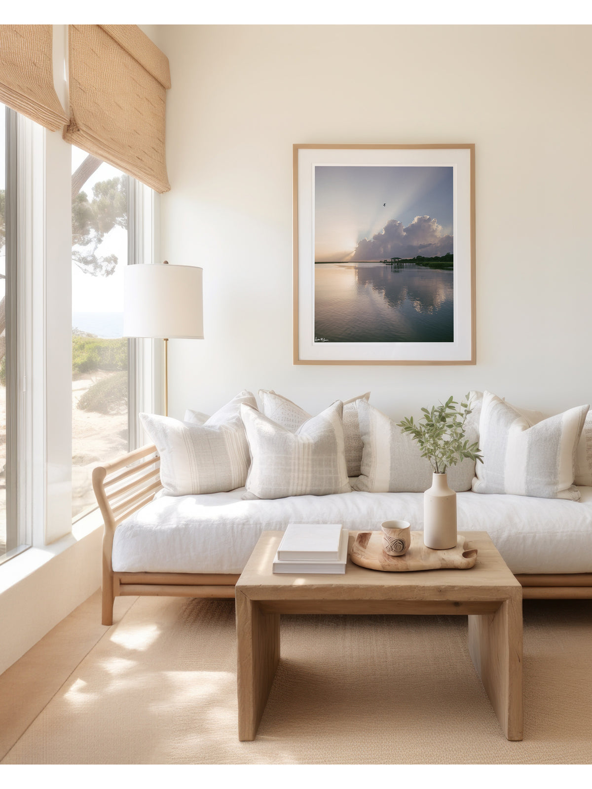 A framed scenic landscape photograph of a marsh during sunrise with sun rays breaking through clouds, a small bird in the sky, and wooden docks extending into the water. The scene is reflected clearly in the still water, with green marsh grass visible along the right shoreline. “Morning Glory” fine art photography print photographed by Kristen M. Brown, Samba to the Sea. Framed print hangs on a cream wall above a light wood sofa with white pillows in a bright, coastal living room.