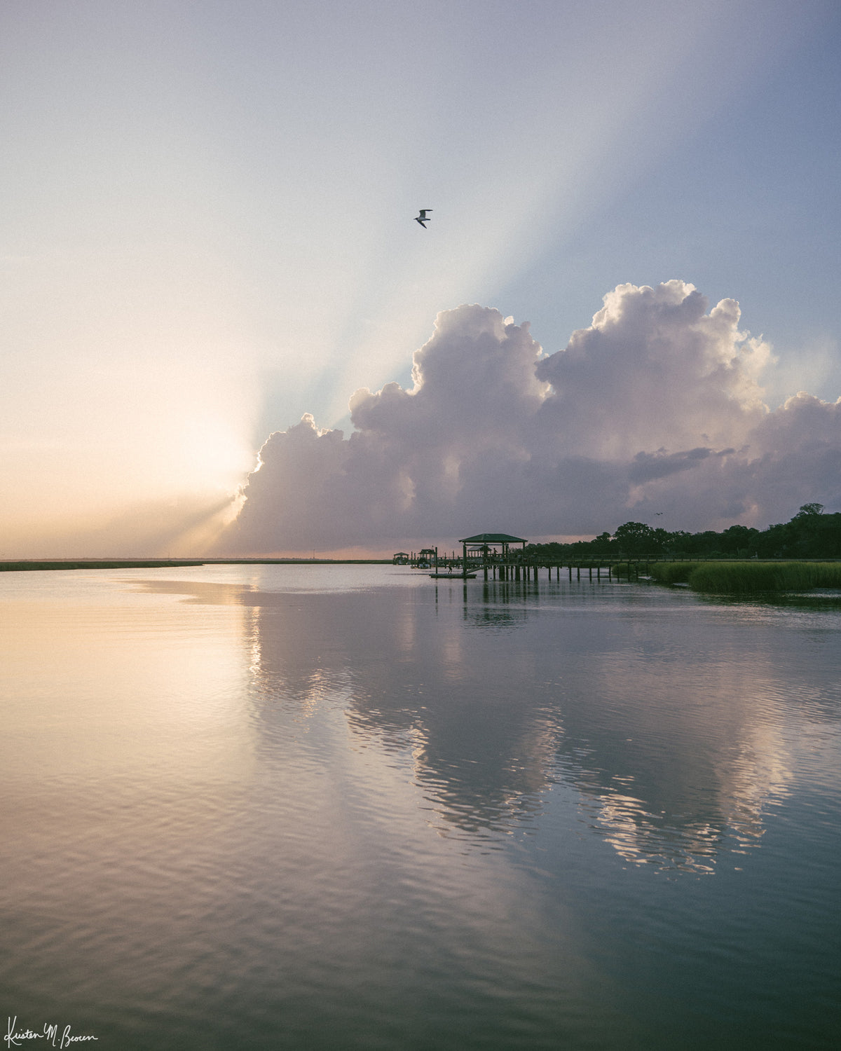 A scenic landscape photograph of a marsh during sunrise with sun rays breaking through clouds, a small bird in the sky, and wooden docks extending into the water. The scene is reflected clearly in the still water, with green marsh grass visible along the right shoreline. “Morning Glory” fine art photography print photographed by Kristen M. Brown, Samba to the Sea.
