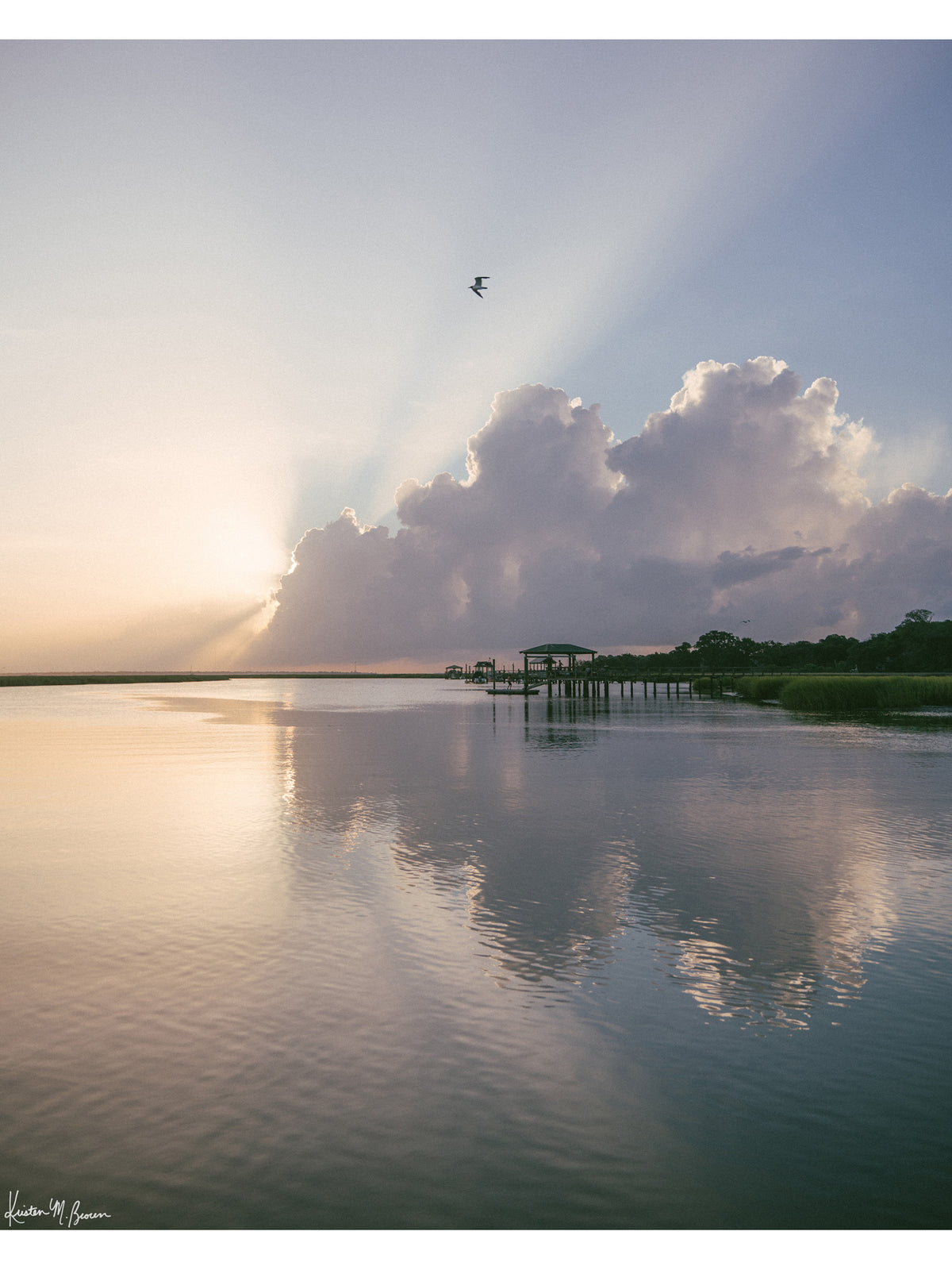A scenic landscape photograph of a marsh during sunrise with sun rays breaking through clouds, a small bird in the sky, and wooden docks extending into the water. The scene is reflected clearly in the still water, with green marsh grass visible along the right shoreline. “Morning Glory” fine art photography print photographed by Kristen M. Brown, Samba to the Sea.
