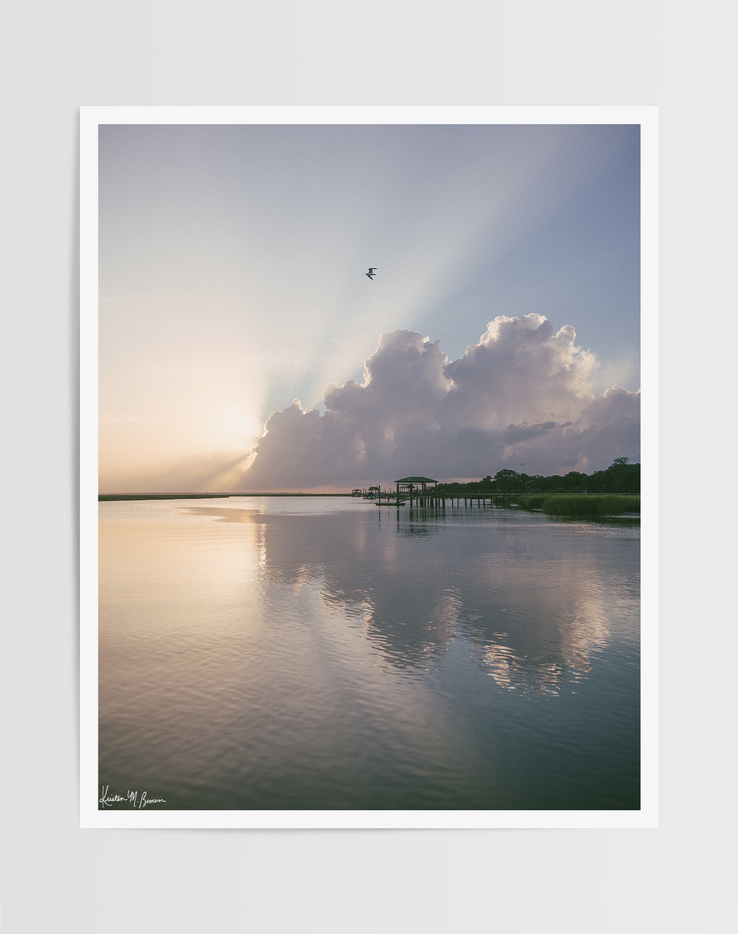 A scenic landscape photograph of a marsh during sunrise with sun rays breaking through clouds, a small bird in the sky, and wooden docks extending into the water. The scene is reflected clearly in the still water, with green marsh grass visible along the right shoreline. “Morning Glory” fine art photography print photographed by Kristen M. Brown, Samba to the Sea.