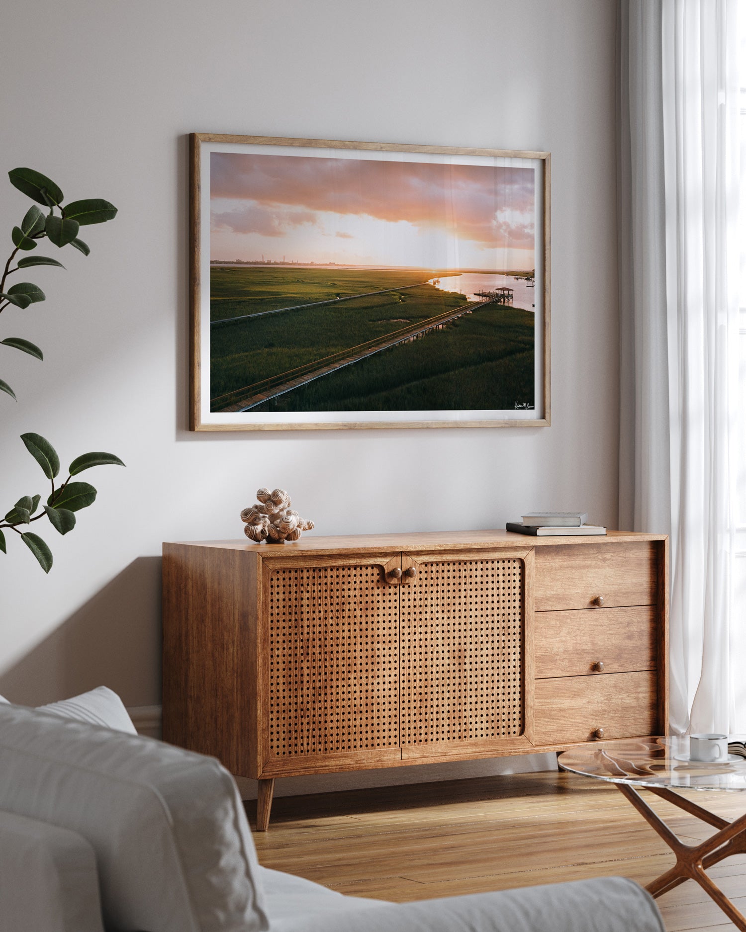 Aerial photograph of pastel pink sunrise over Charleston harbor and marsh with a boardwalk path, waterway, and view of downtown Charleston in the background. "Morning Harbor View" fine art photography print photographed by Kristen M. Brown, Samba to the Sea. Frame print displayed on a light gray wall above a wooden credenza in a modern living room setting.
