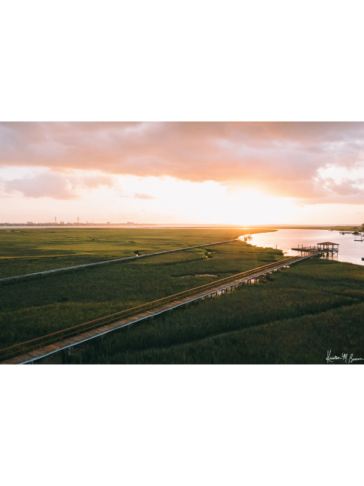Aerial photograph of pastel pink sunrise over Charleston harbor and marsh with a boardwalk path, waterway, and view of downtown Charleston in the background. "Morning Harbor View" fine art photography print photographed by Kristen M. Brown, Samba to the Sea.
