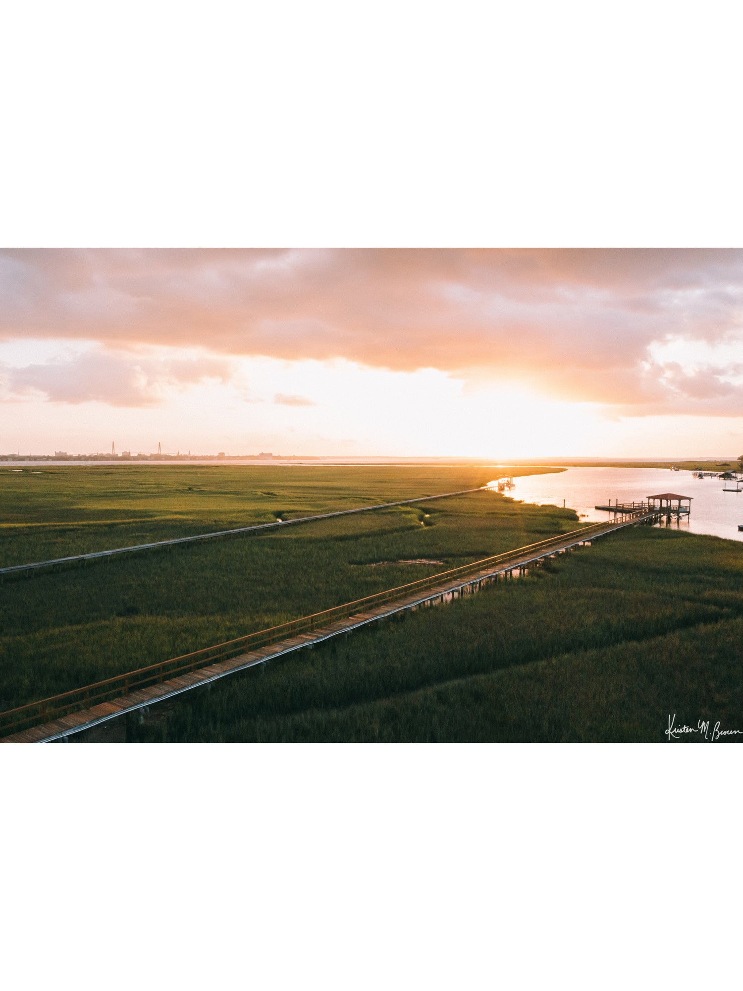 Aerial photograph of pastel pink sunrise over Charleston harbor and marsh with a boardwalk path, waterway, and view of downtown Charleston in the background. "Morning Harbor View" fine art photography print photographed by Kristen M. Brown, Samba to the Sea.