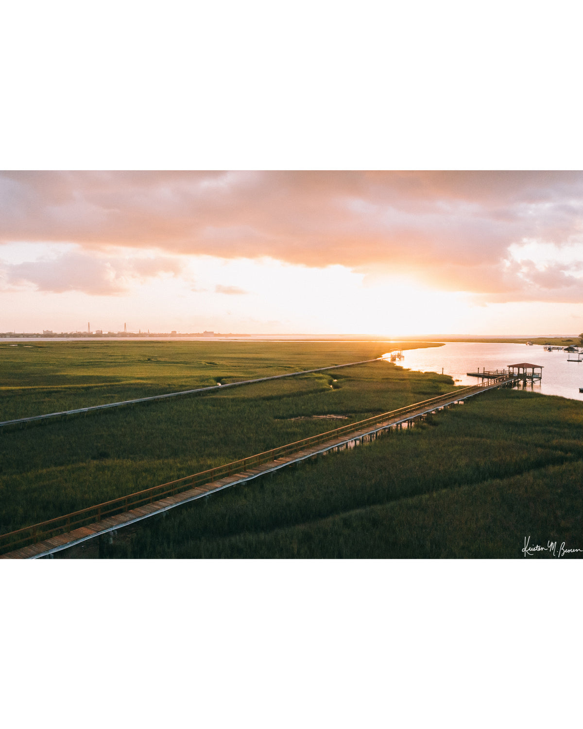 Aerial photograph of pastel pink sunrise over Charleston harbor and marsh with a boardwalk path, waterway, and view of downtown Charleston in the background. "Morning Harbor View" fine art photography print photographed by Kristen M. Brown, Samba to the Sea.