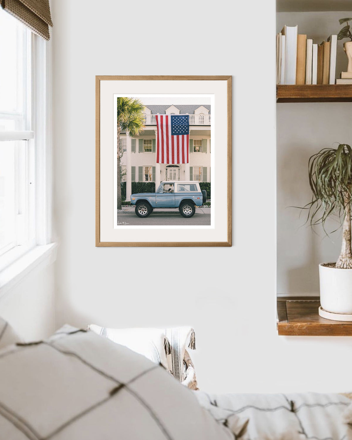 Framed photo print of a dog sitting in the driver's seat of a light blue vintage Ford Bronco parked on the street in front of a large white colonial-style house with an oversized American flag hanging from the front porch in Charleston, SC. Murphy's driving, are you calling shotgun? Hop on in, enjoy the ride, and bring home that feeling of cruising around in one gorgeous vintage Bronco, windows down, and wind flowing through your hair. Photographed by Kristen M. Brown for The Sunset Shop.