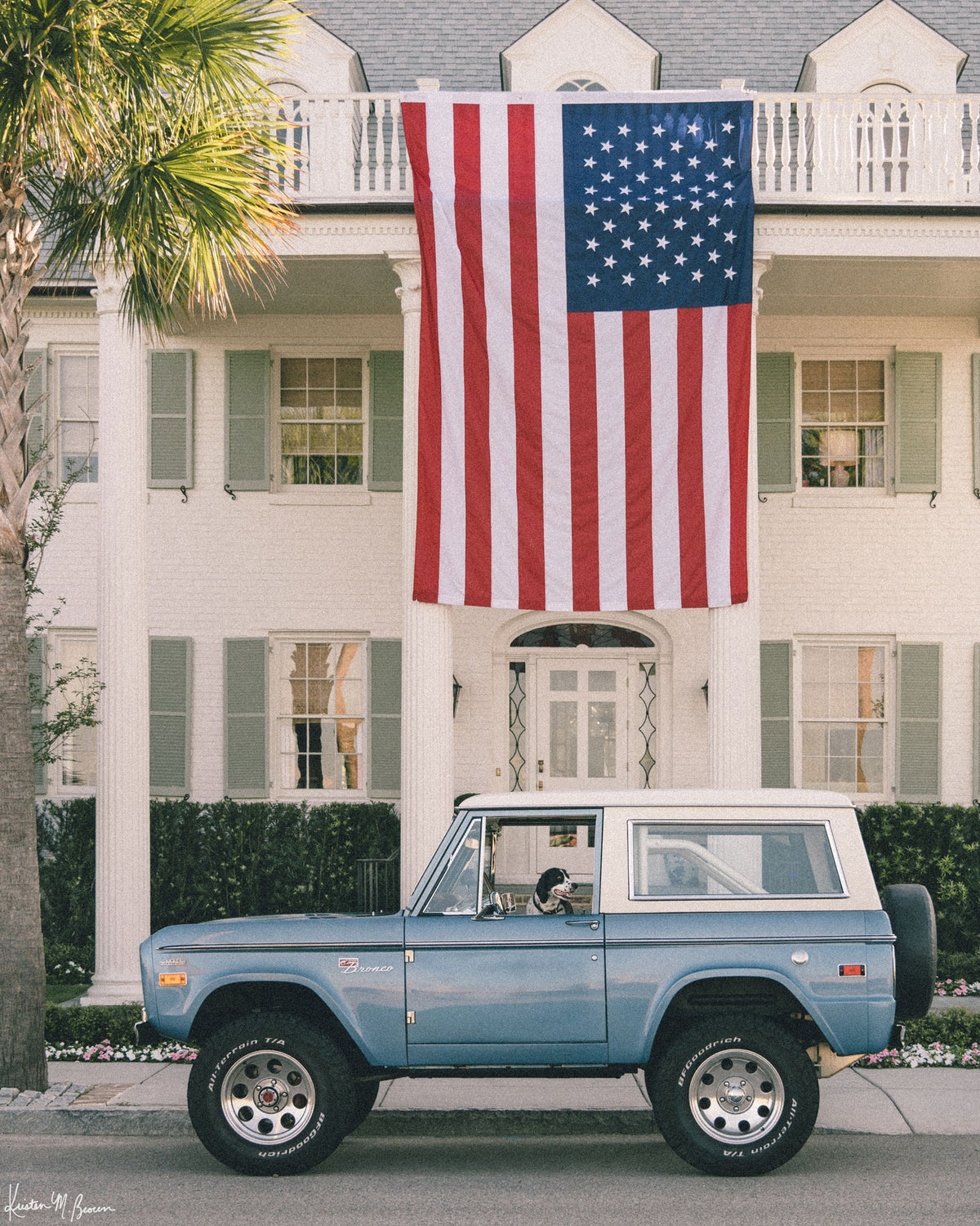 Photo print of a dog sitting in the driver's seat of a light blue vintage Ford Bronco parked on the street in front of a large white colonial-style house with an oversized American flag hanging from the front porch in Charleston, SC. Murphy's driving, are you calling shotgun? Hop on in, enjoy the ride, and bring home that feeling of cruising around in one gorgeous vintage Bronco, windows down, and wind flowing through your hair. Photographed by Kristen M. Brown for The Sunset Shop.