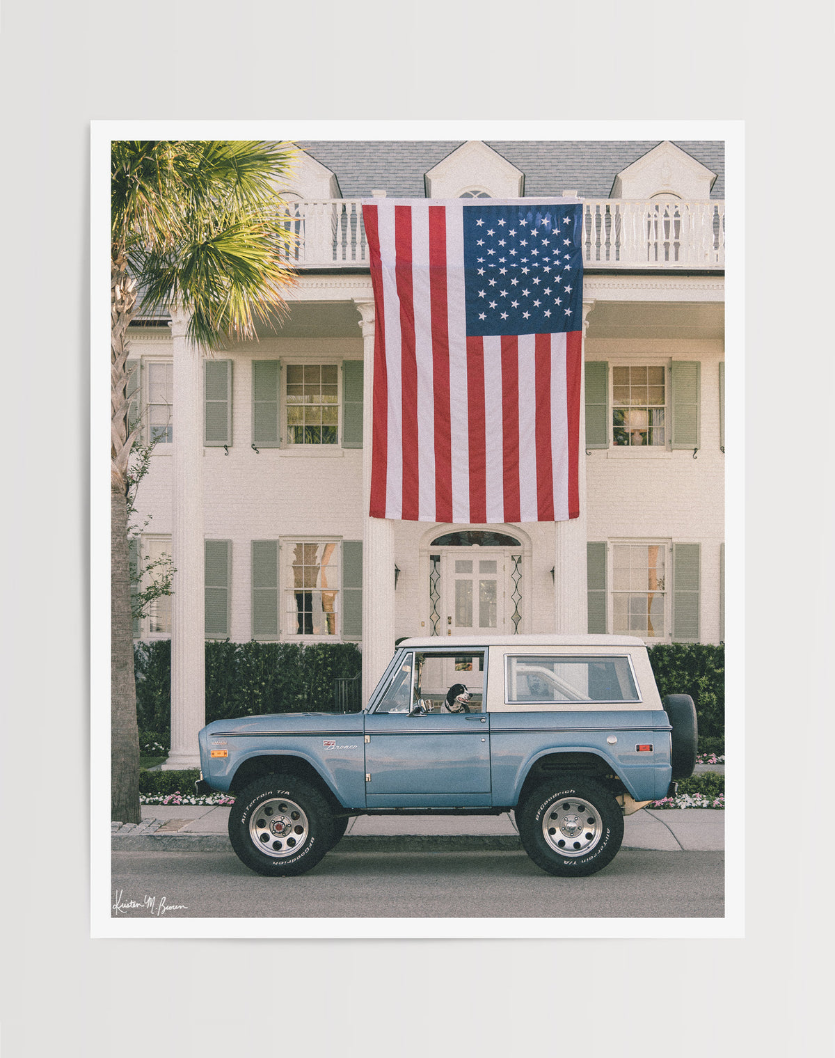 Photo print of a dog sitting in the driver's seat of a light blue vintage Ford Bronco parked on the street in front of a large white colonial-style house with an oversized American flag hanging from the front porch in Charleston, SC. Murphy's driving, are you calling shotgun? Hop on in, enjoy the ride, and bring home that feeling of cruising around in one gorgeous vintage Bronco, windows down, and wind flowing through your hair. Photographed by Kristen M. Brown for The Sunset Shop.