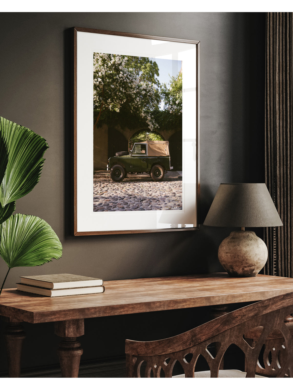A framed art print featuring a vintage Land Rover series truck print, styled on a dark wall above a wooden console table with a lamp and books. A vintage green Land Rover Series truck British Army version with tan canvas top, parked on a cobblestone street in historic Charleston with blooming white Crepe Myrtle flowers. “Passage to Chalmers” is a journey into the charming streets of historic Charleston, where beauty and nostalgia meet. Vintage Land Rover photo print by Kristen M. Brown of Samba to the Sea.