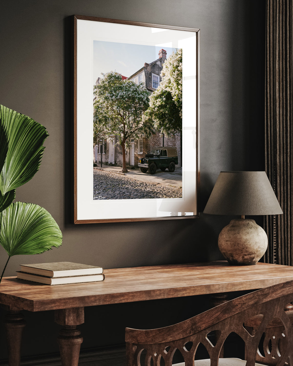A framed vintage Land Rover photo print, displayed on a dark gray wall above a wooden console table with a lamp and books. A vintage green Land Rover Series truck British Army version with tan canvas top, parked on a cobblestone street in historic Charleston with blooming white Crepe Myrtle flowers. “Rover Blooms” is a journey into the charming streets of historic Charleston, where beauty and nostalgia meet. Vintage Land Rover photo print by Kristen M. Brown of Samba to the Sea for The Sunset Shop.