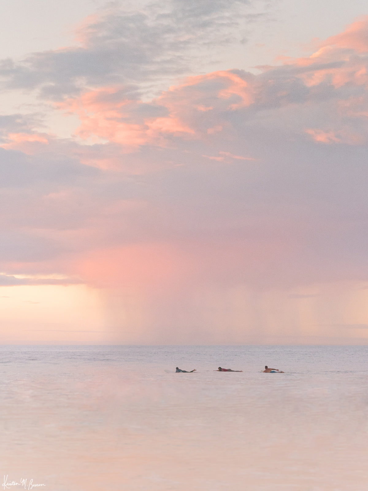 Surfers paddling in the ocean during a pastel pink rain squall at sunset in Costa Rica. What a surfer&#39;s dreams are made of - - beautiful sunset surf sessions. Have a piece of sunset surfer magic in your very own home with &quot;Saltwater Paradise&quot; sunset surfer wall art photographed by Kristen M. Brown of Samba to the Sea for The Sunset Shop.