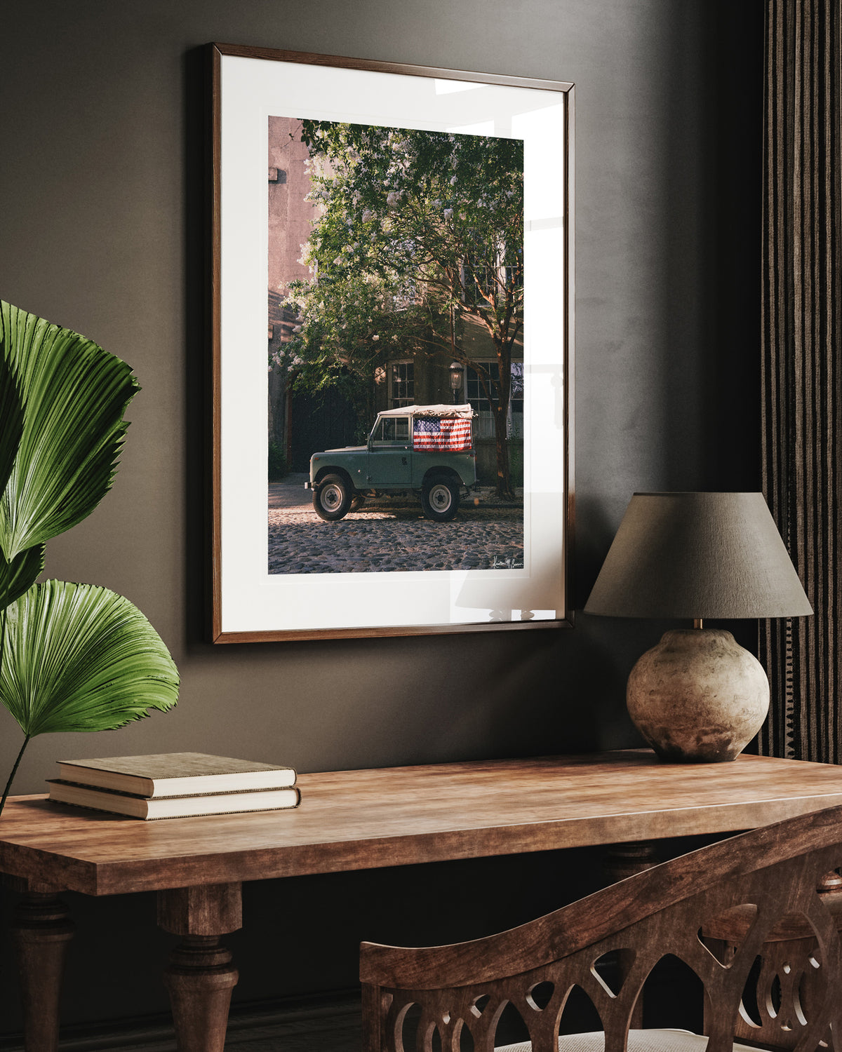 Framed art print featuring vintage green Land Rover Series with an American flag draped over the rear window, parked on a cobblestone street in historic Charleston with blooming white Crepe Myrtle flowers, displayed on a dark wall above a wooden console table with a lamp and books. "Stars & Stripes & Series" is a journey into the charming streets of historic Charleston, where beauty and nostalgia meet. Vintage Land Rover photo print by Kristen M. Brown of Samba to the Sea for The Sunset Shop.