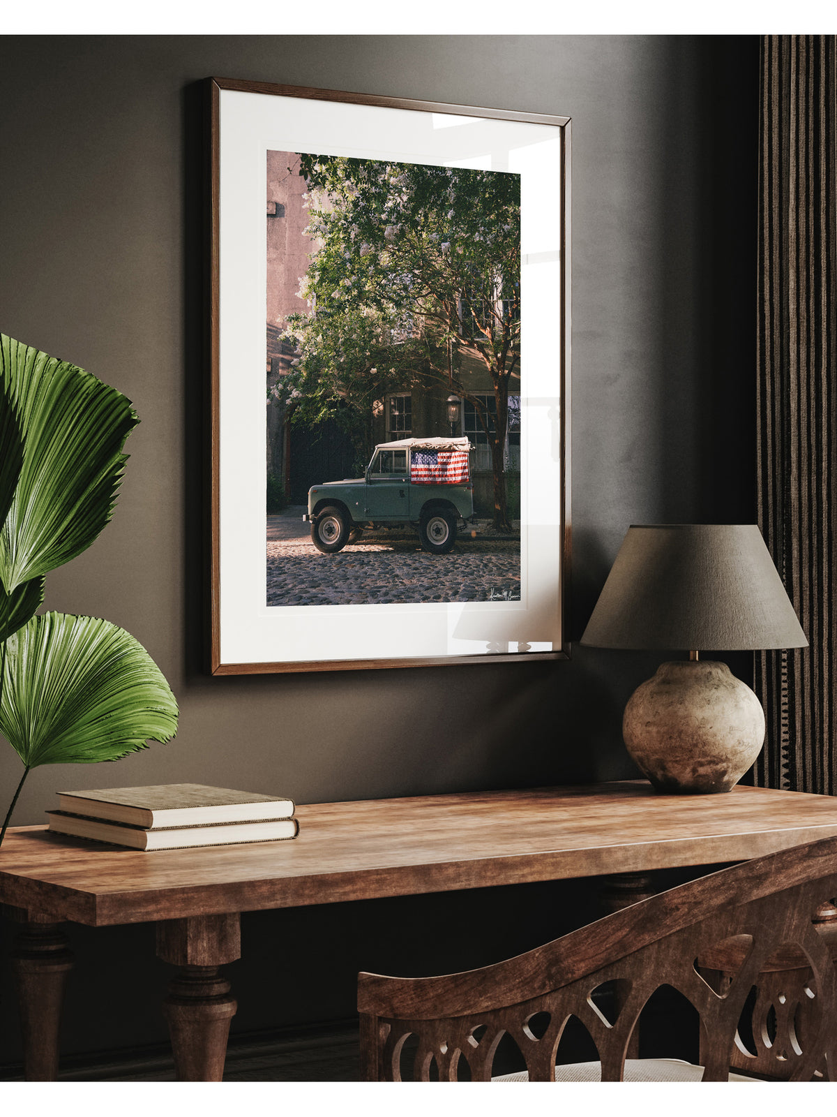 Framed art print featuring vintage green Land Rover Series with an American flag draped over the rear window, parked on a cobblestone street in historic Charleston with blooming white Crepe Myrtle flowers, displayed on a dark wall above a wooden console table with a lamp and books. "Stars & Stripes & Series" is a journey into the charming streets of historic Charleston, where beauty and nostalgia meet. Vintage Land Rover photo print by Kristen M. Brown of Samba to the Sea for The Sunset Shop.