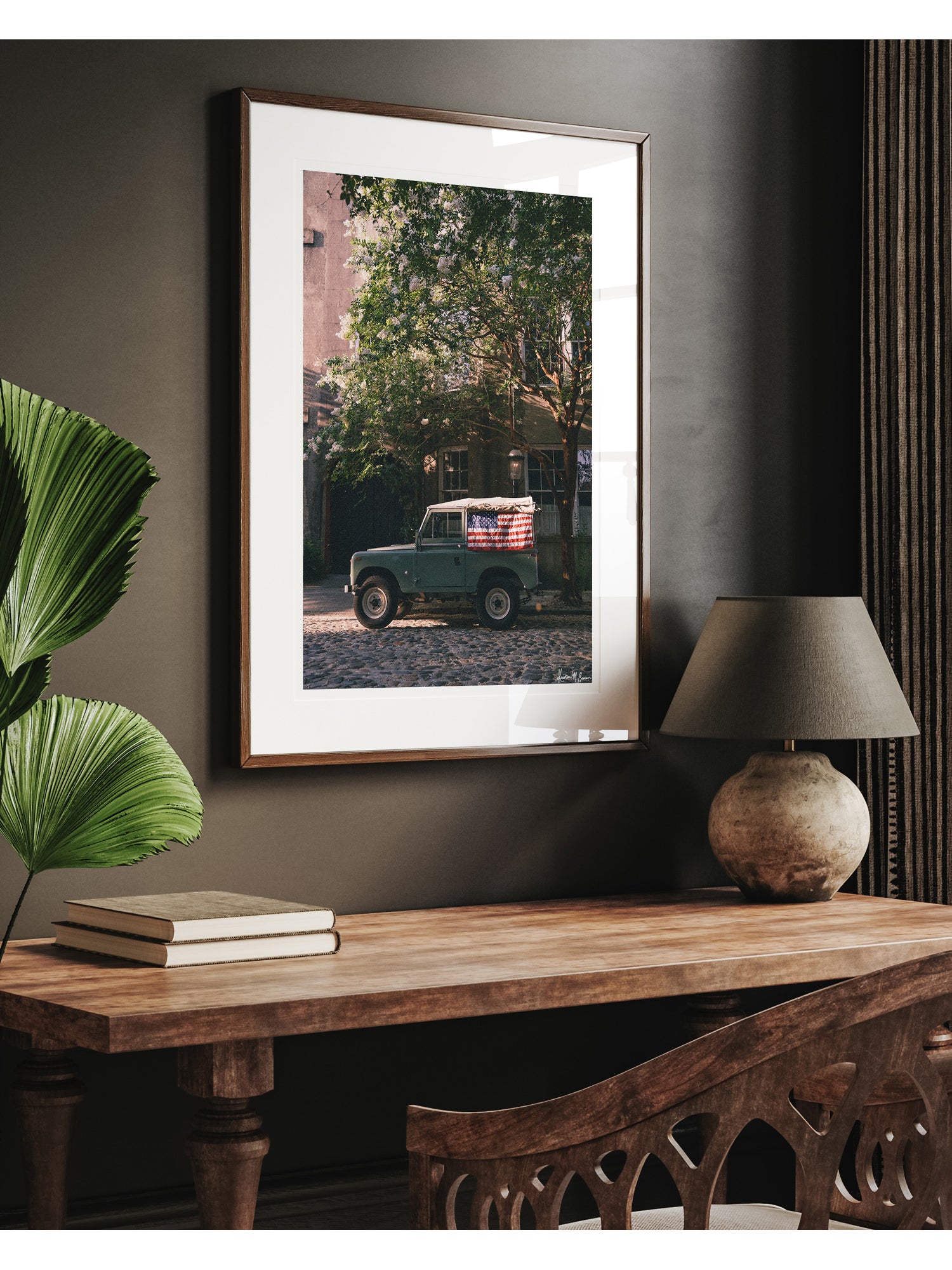 Framed art print featuring vintage green Land Rover Series with an American flag draped over the rear window, parked on a cobblestone street in historic Charleston with blooming white Crepe Myrtle flowers, displayed on a dark wall above a wooden console table with a lamp and books. "Stars & Stripes & Series" is a journey into the charming streets of historic Charleston, where beauty and nostalgia meet. Vintage Land Rover photo print by Kristen M. Brown of Samba to the Sea for The Sunset Shop.